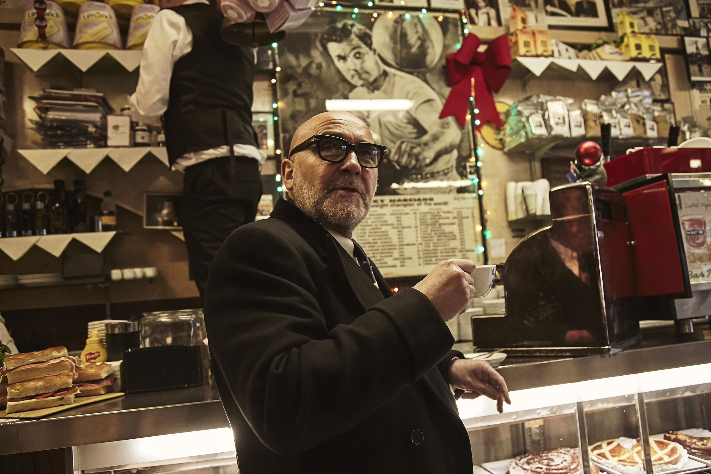 An elderly man in a black suit, glasses, and with a beard is sitting at a counter in a diner, holding a small cup of coffee. Behind him, there is a person wearing a black vest and white shirt, with their back to the camera. The wall in the background