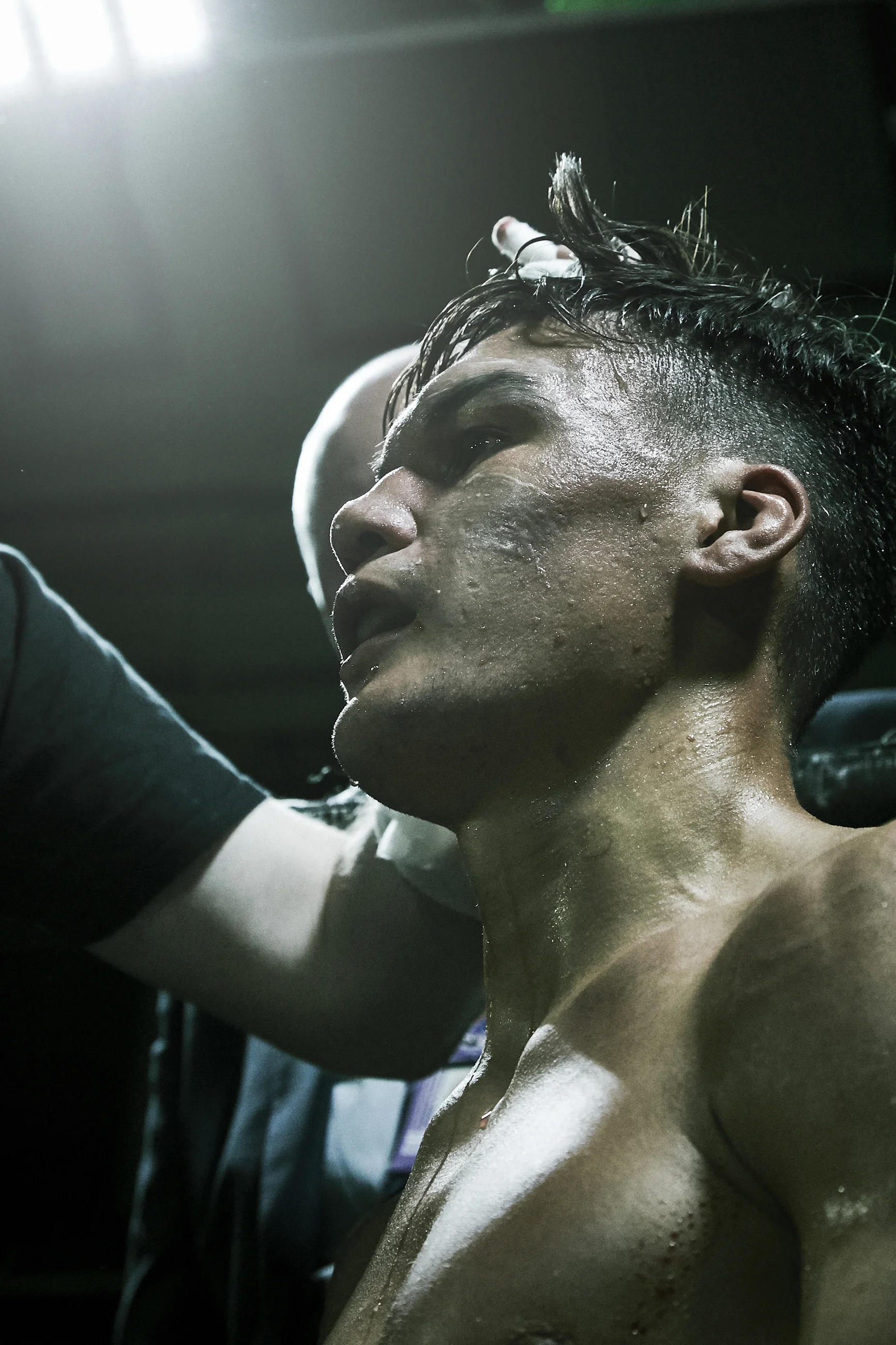 A shirtless male athlete with wet hair, sweat, and dirt on his face, looking exhausted, receiving medical attention from a person wearing gloves.