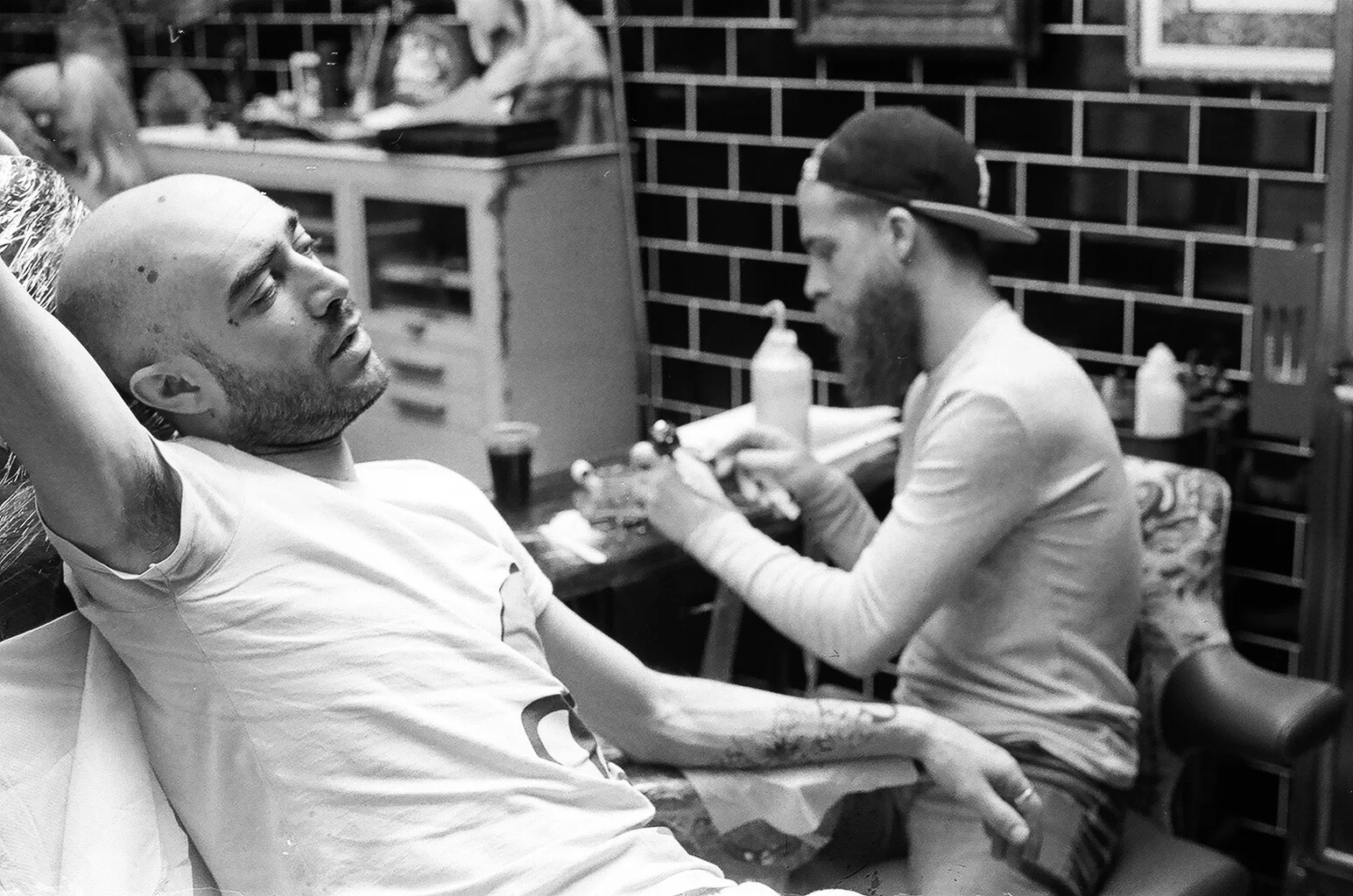 A man with a bald head and tattoos on his arm is lying on a medical chair, receiving a tattoo. He appears relaxed. In the background, a tattoo artist with a beard, wearing a backward cap and gloves, is working on another tattoo. The setting is a tatt