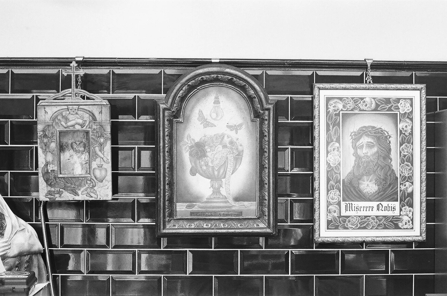 Three framed religious or spiritual prints hanging on a dark tiled wall. The left print shows a skeleton with a rat and various objects, the middle print depicts a heart in a chalice with butterflies and a snake, and the right print illustrates a bea
