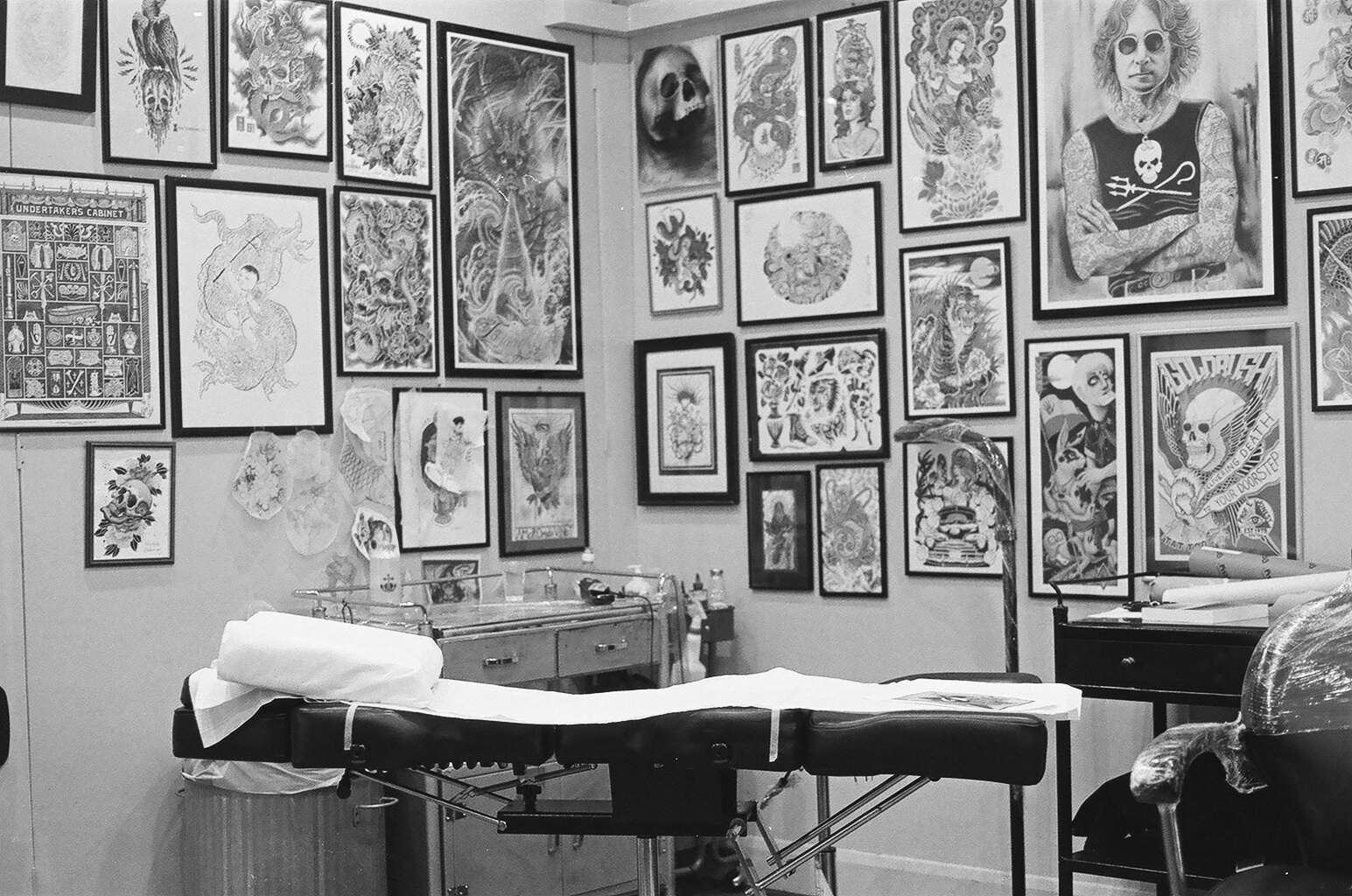 Tattoo studio with tattoo chair in foreground, walls decorated with numerous tattoo artwork and posters, including skulls, dragons, and portraits, along with a tattooing station and supplies.