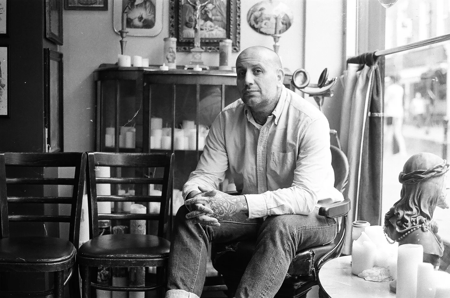 A man with a shaved head and tattoos on his right arm, sitting on a chair in a room with various decorations, candles, and a sculpture of a woman with a headscarf, next to a window.