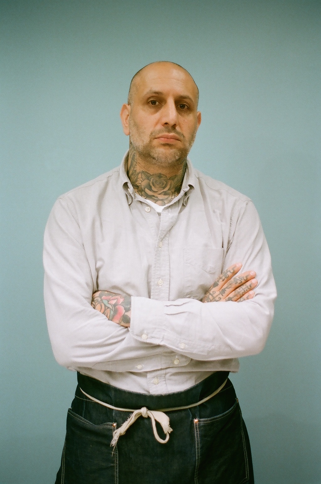 A bald man with tattoos on his neck and hands, wearing a light-colored button-up shirt and black pants, standing with arms crossed against a plain blue background.