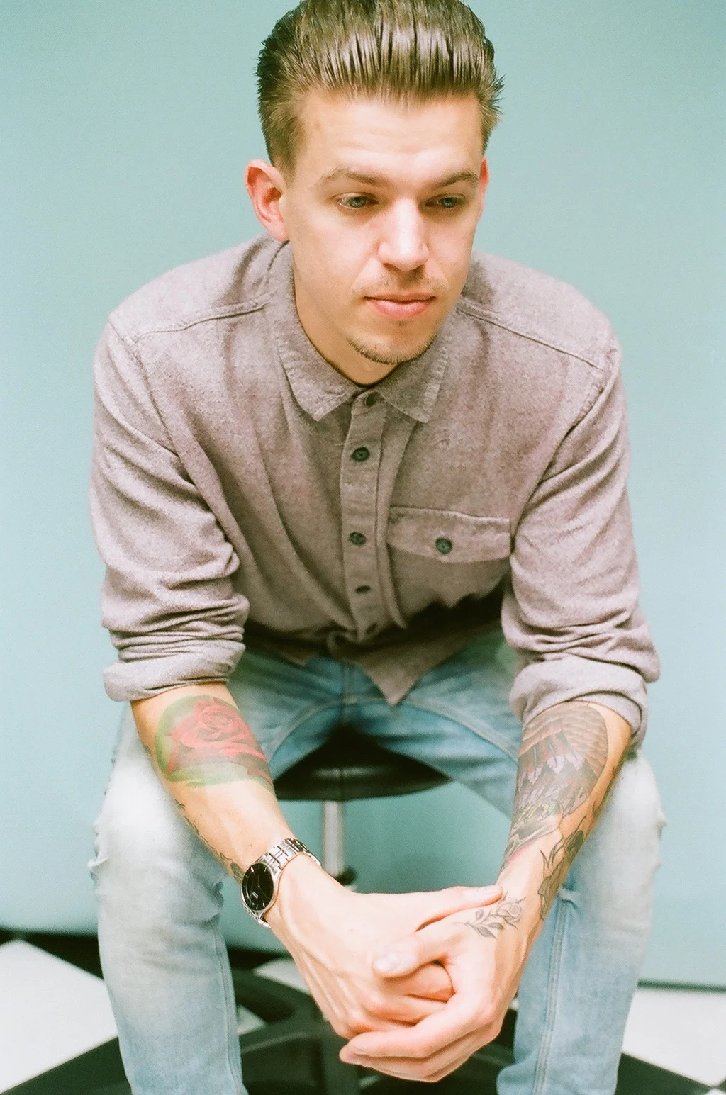 A man with tattoos on his arms, wearing a gray button-up shirt and a watch, is sitting on a stool with his hands clasped, against a teal background.