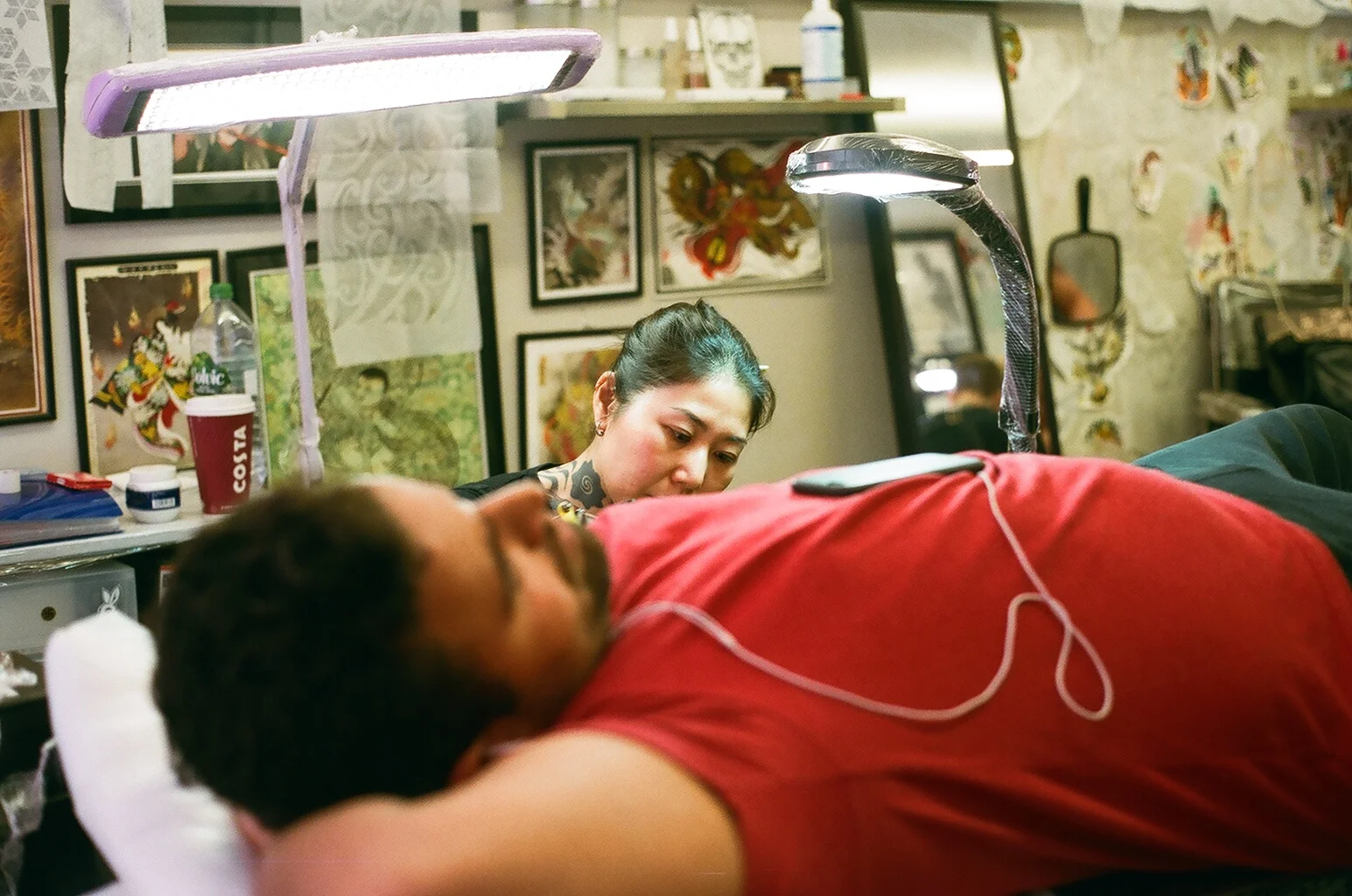 A woman getting a tattoo on a person's side at an art studio, with tattoo artwork and framed pictures on the wall behind them, and various tattoo supplies visible.