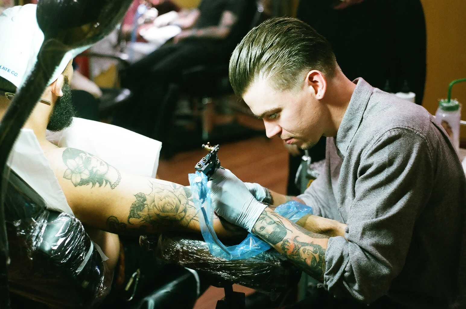 A tattoo artist with short, slicked-back hair is tattooing a person's arm in a tattoo studio. The artist is wearing gloves, focusing on his work, and has colorful tattoos on his arms. The person's arm being tattooed already has multiple tattoos, incl