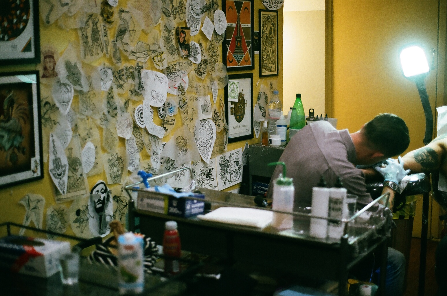 Tattoo artist working on a client's arm in a tattoo studio with yellow walls decorated with various tattoo designs and art.