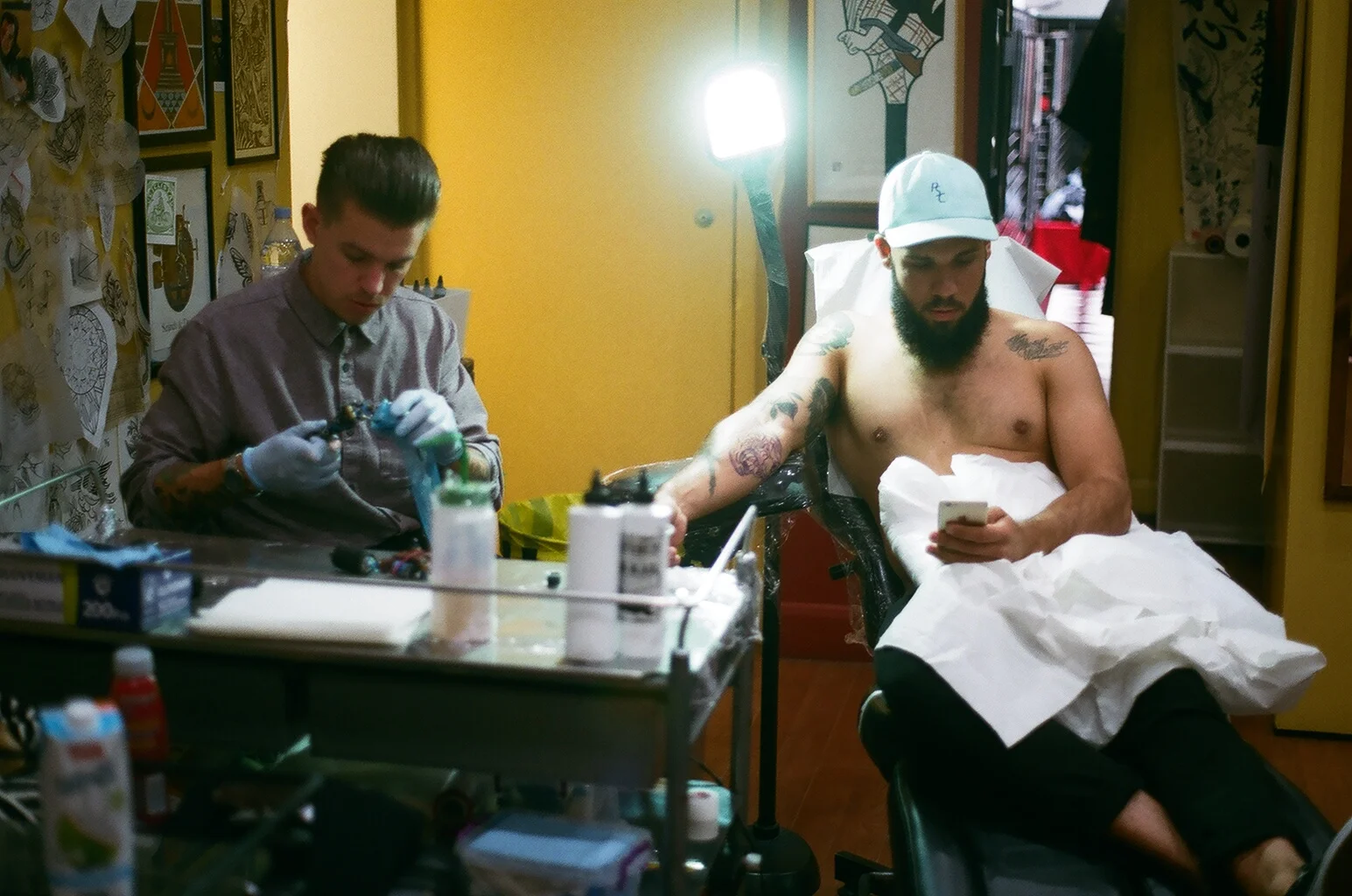 A man receiving a tattoo from a tattoo artist in a studio, sitting shirtless on a chair with his phone, while the tattoo artist is working on his arm.