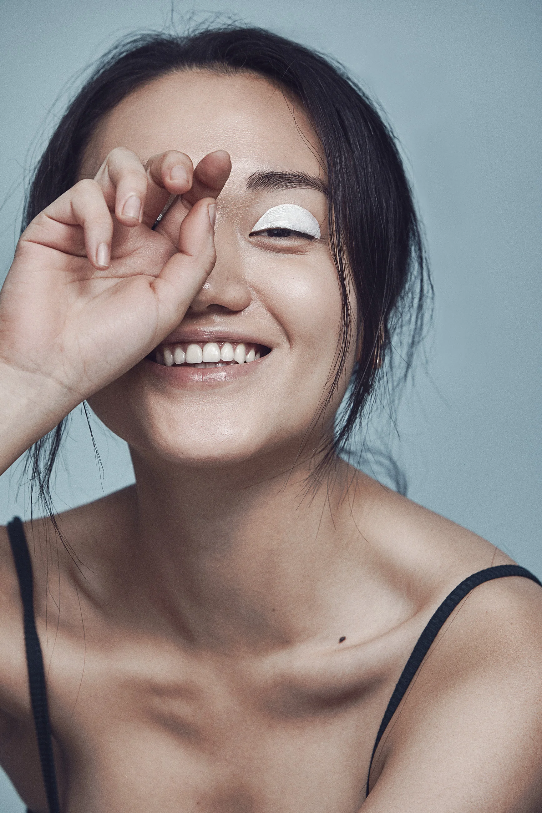 A woman smiling and looking at the camera, with her hand near her face and eye makeup in white on her eyelid, wearing a black spaghetti strap top.