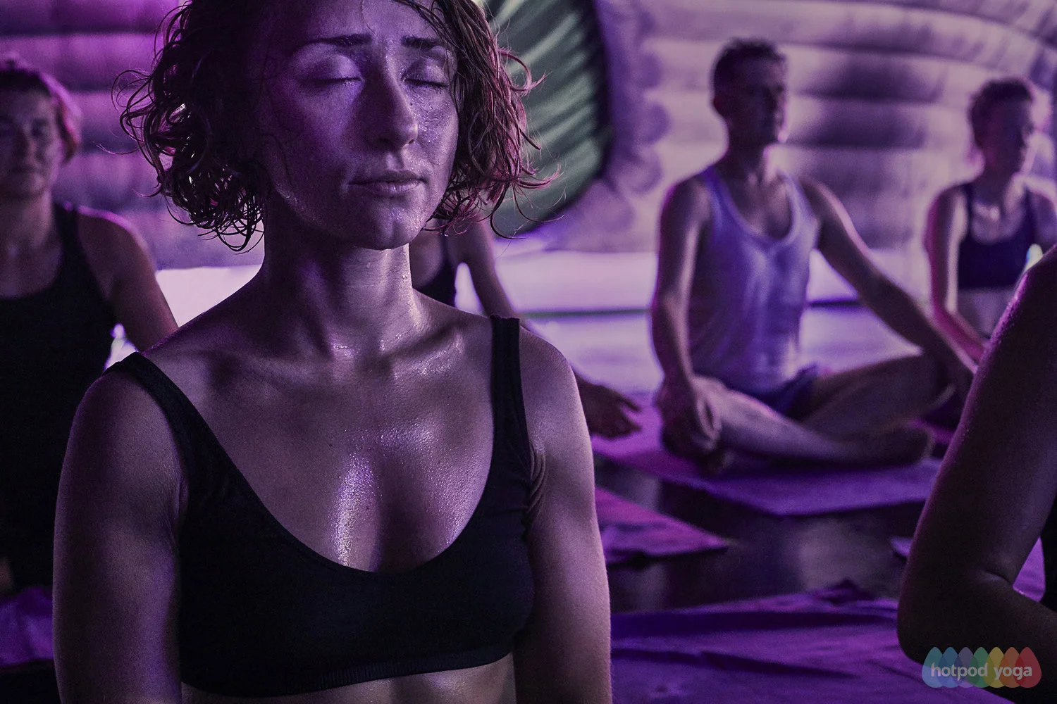 A woman with wet hair and closed eyes participating in a meditation class among other students in a dimly lit yoga studio.