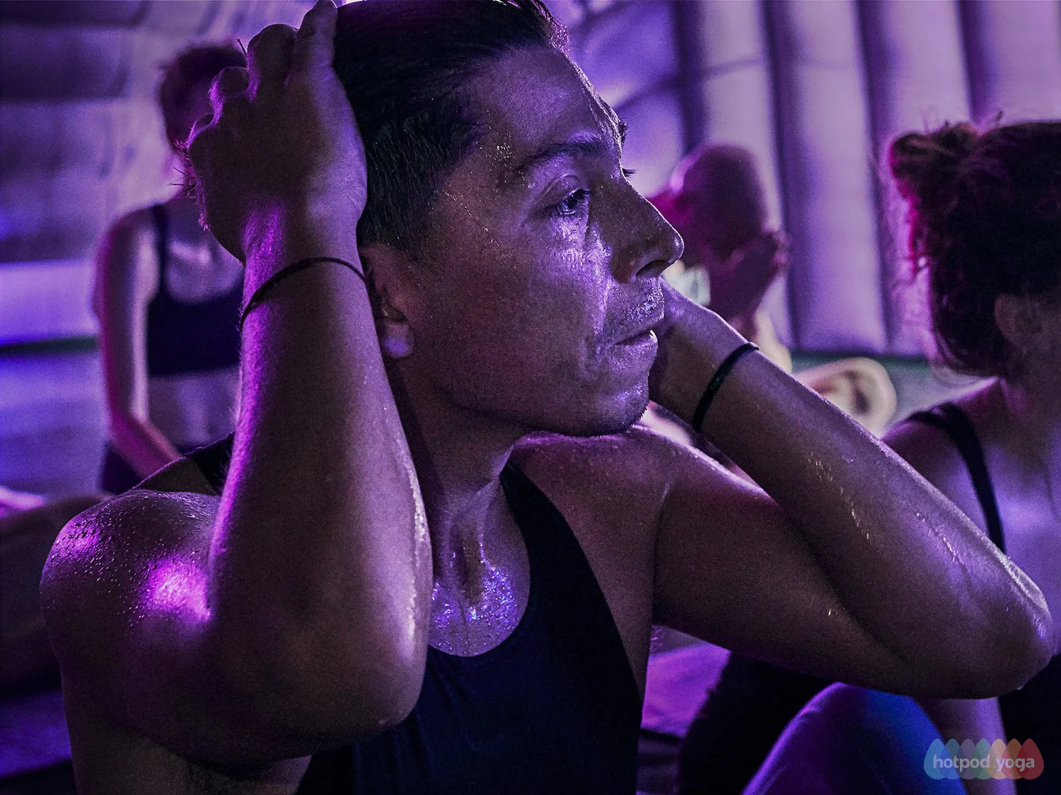 A woman with dark hair in a bun and wearing a black tank top is practicing yoga, sitting against a wall with her hands on her head. She appears to be focused, with her eyes closed and water or sweat on her skin. Other people are doing yoga in the bac