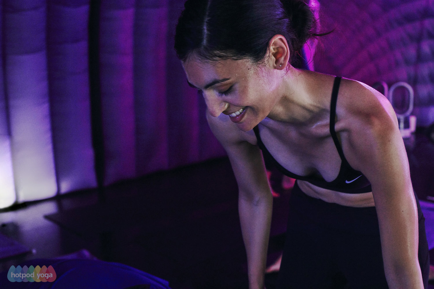 A woman in a sports bra smiling, leaning forward in a yoga or fitness studio with purple lighting.