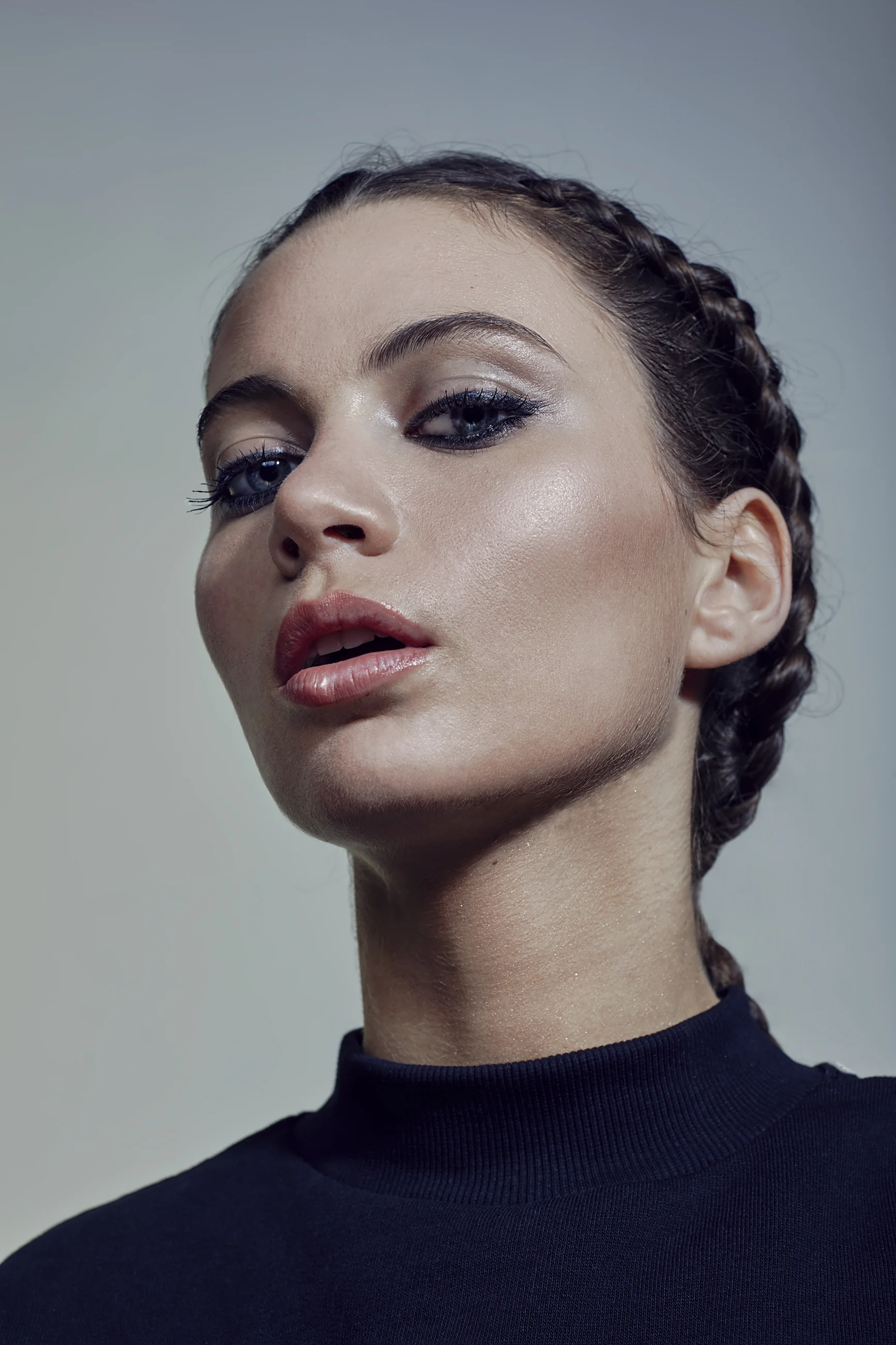 A close-up portrait of a young woman with braided hair, seductive makeup, and a black top, against a gray background.