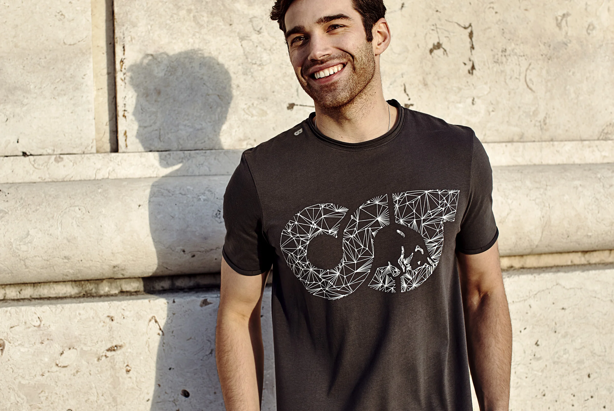 A young man with dark hair, smiling, stands outdoors against a stone wall, wearing a black t-shirt with a geometric graphic design of the letters CDC and a lion's face in the center.