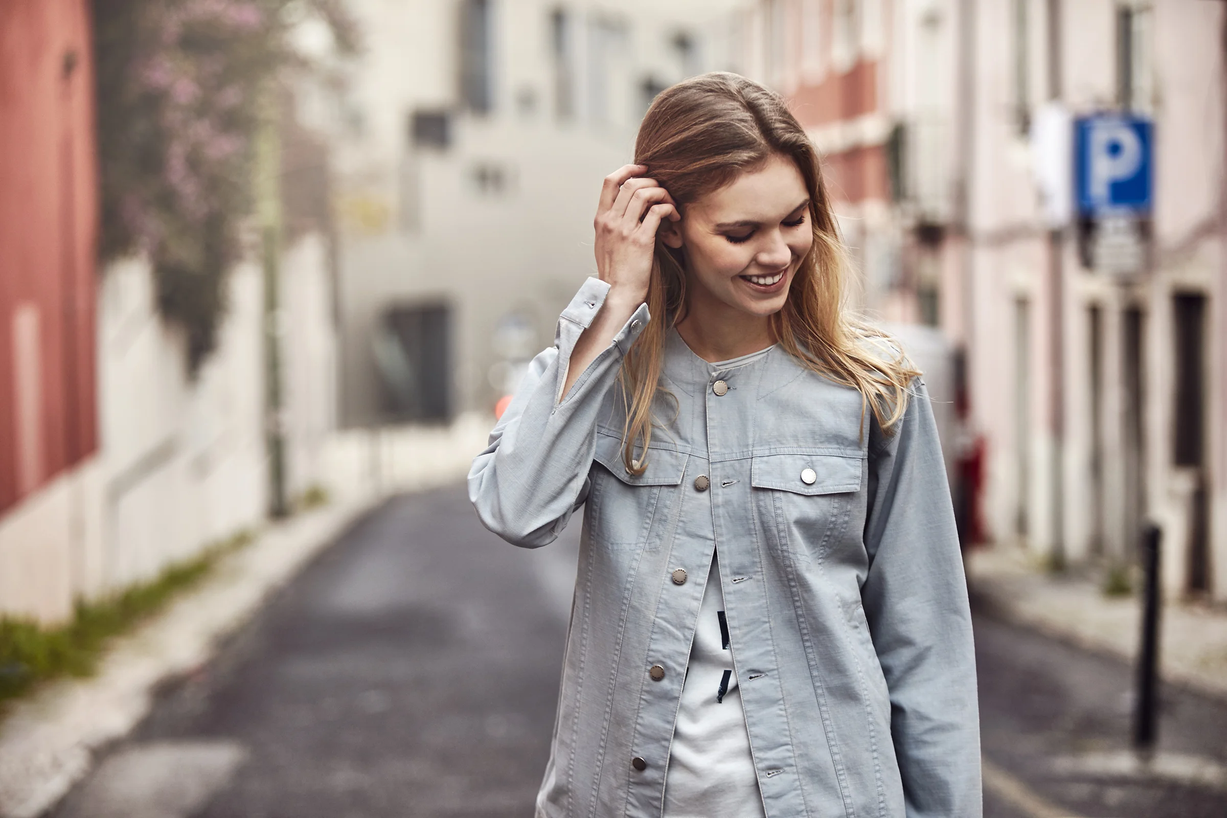 A young woman with long, wavy hair smiling and looking down while walking on a city street, wearing a light gray denim jacket.