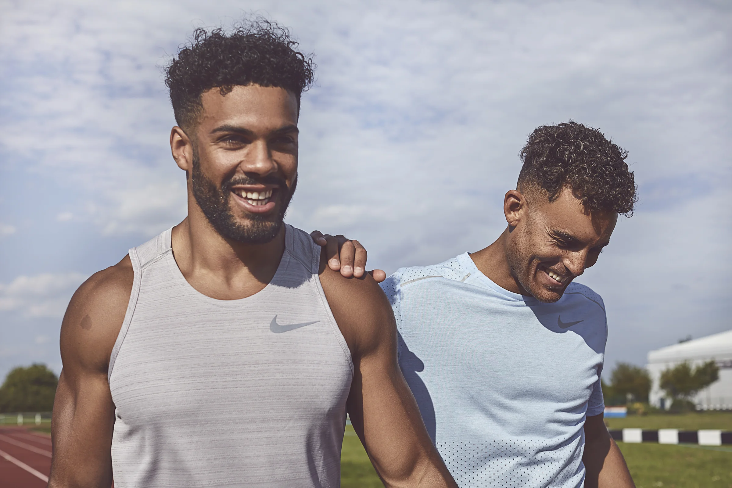 Two male runners smiling at a track field, one resting his hand on the other's shoulder, both wearing running gear.