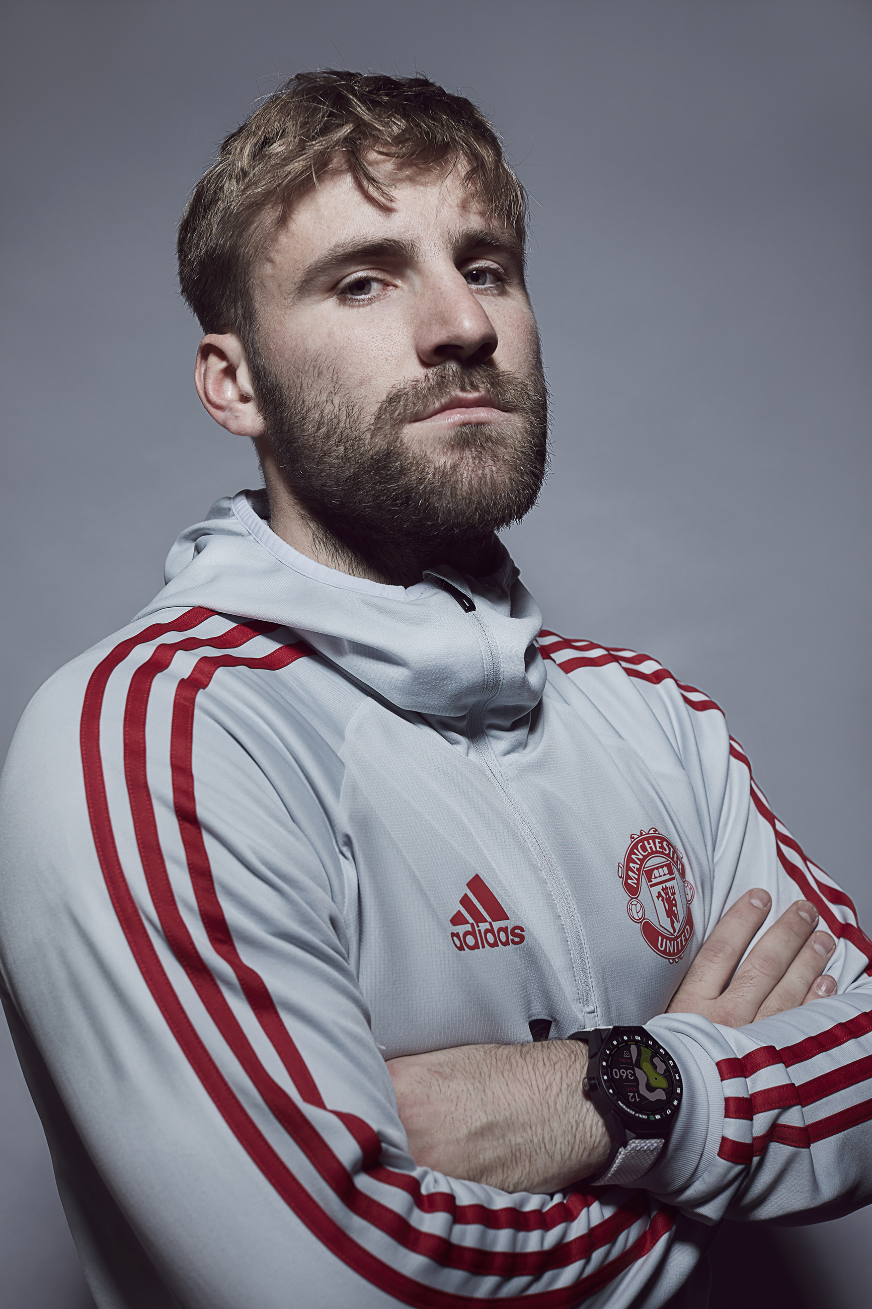 A man with light brown hair and a beard wearing a gray Manchester United athletic jacket with red stripes, crossed arms, and a smartwatch on his left wrist, posing against a plain gray background.