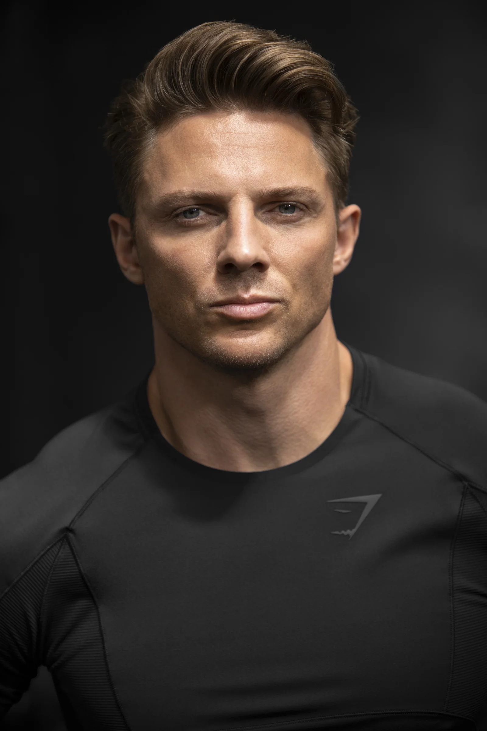 A young man with short, styled brown hair, wearing a black athletic shirt against a dark background.