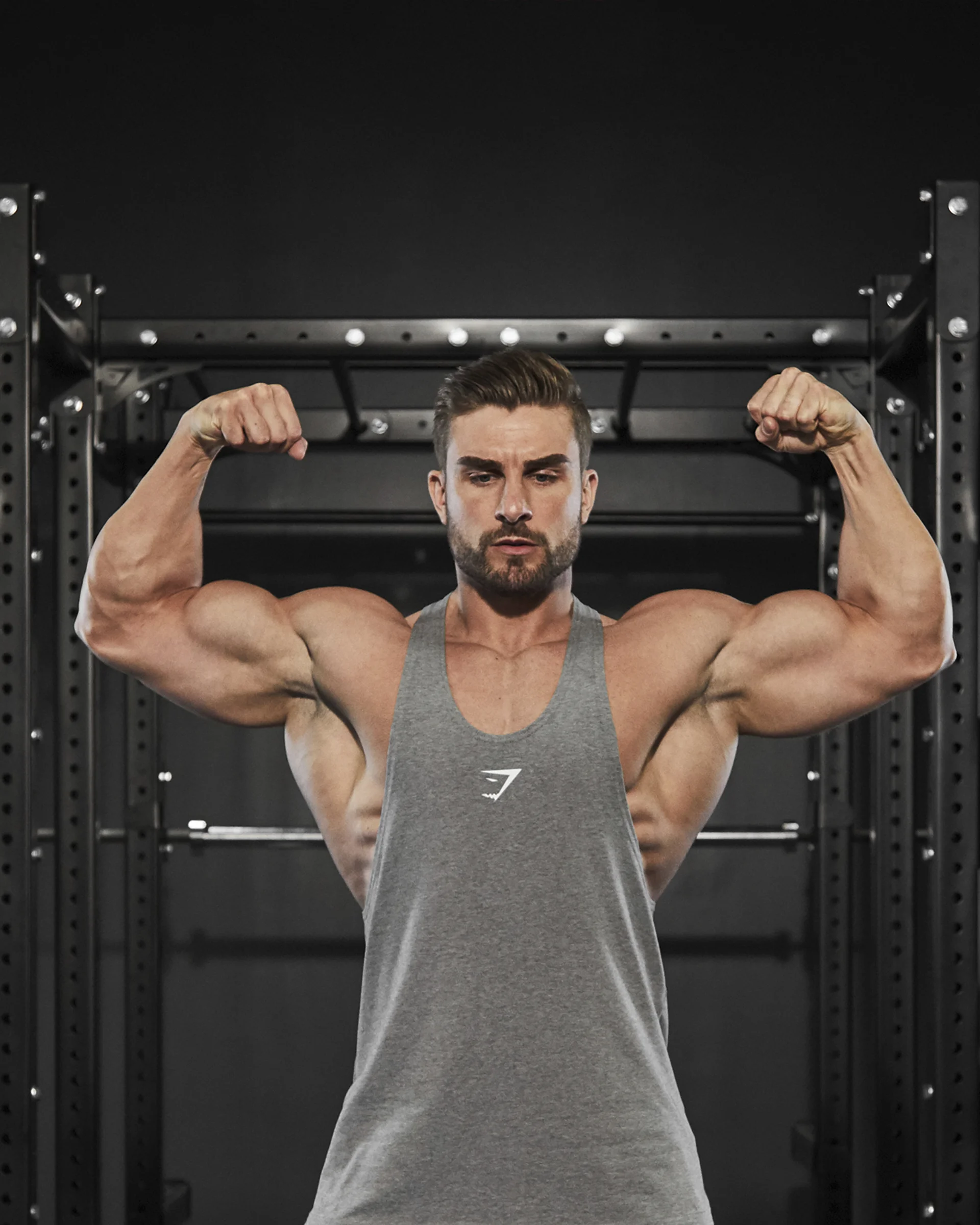 A muscular man flexing his biceps in a gym, wearing a gray tank top.