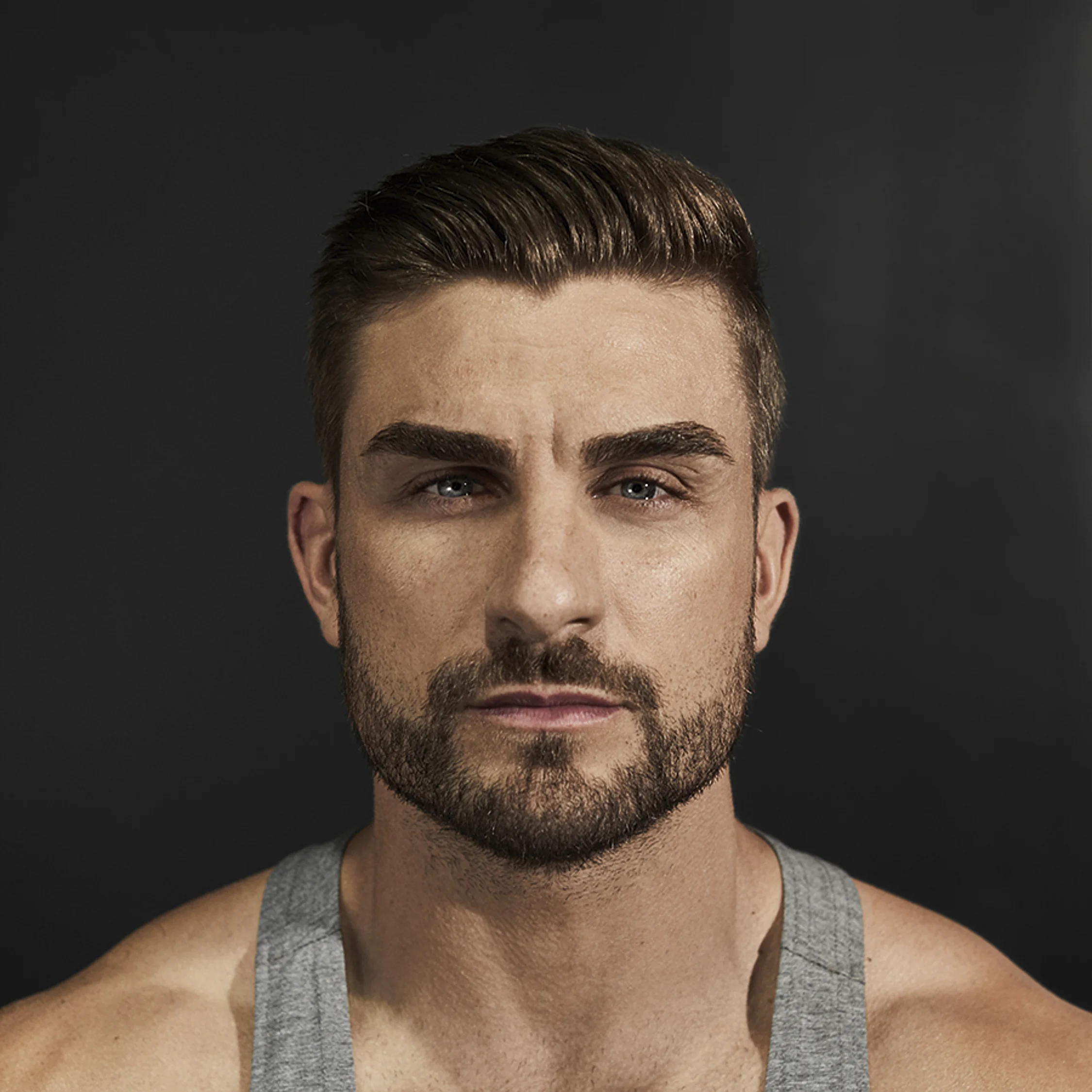 Close-up portrait of a man with styled hair, beard, wearing a gray tank top, against a dark background.