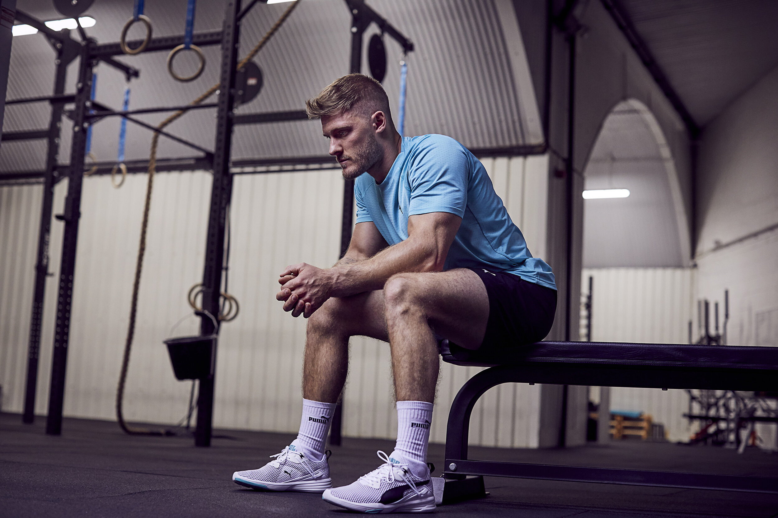A man sitting on a bench in a gym, wearing a blue shirt, black shorts, white Puma socks, and white running shoes, with a workout area and gym equipment in the background.