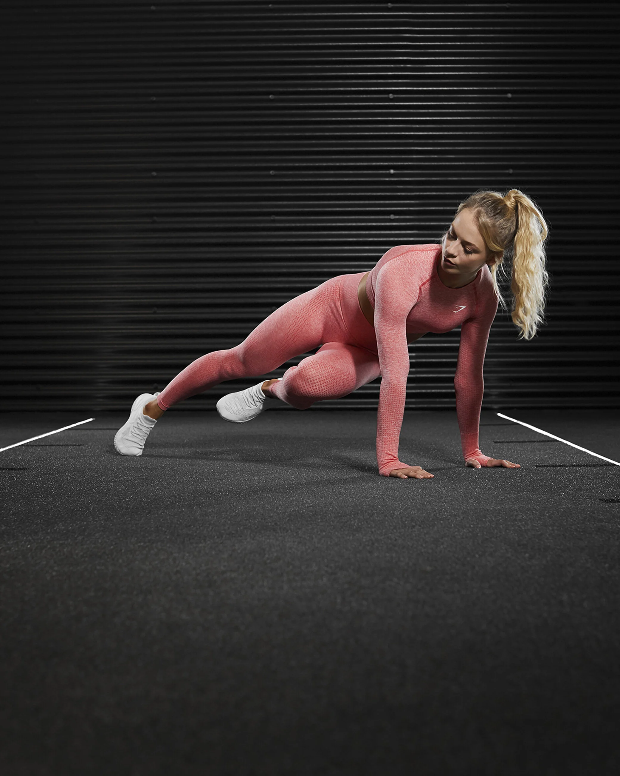 A woman in pink workout attire performing a plank exercise in a gym with a black wall background.