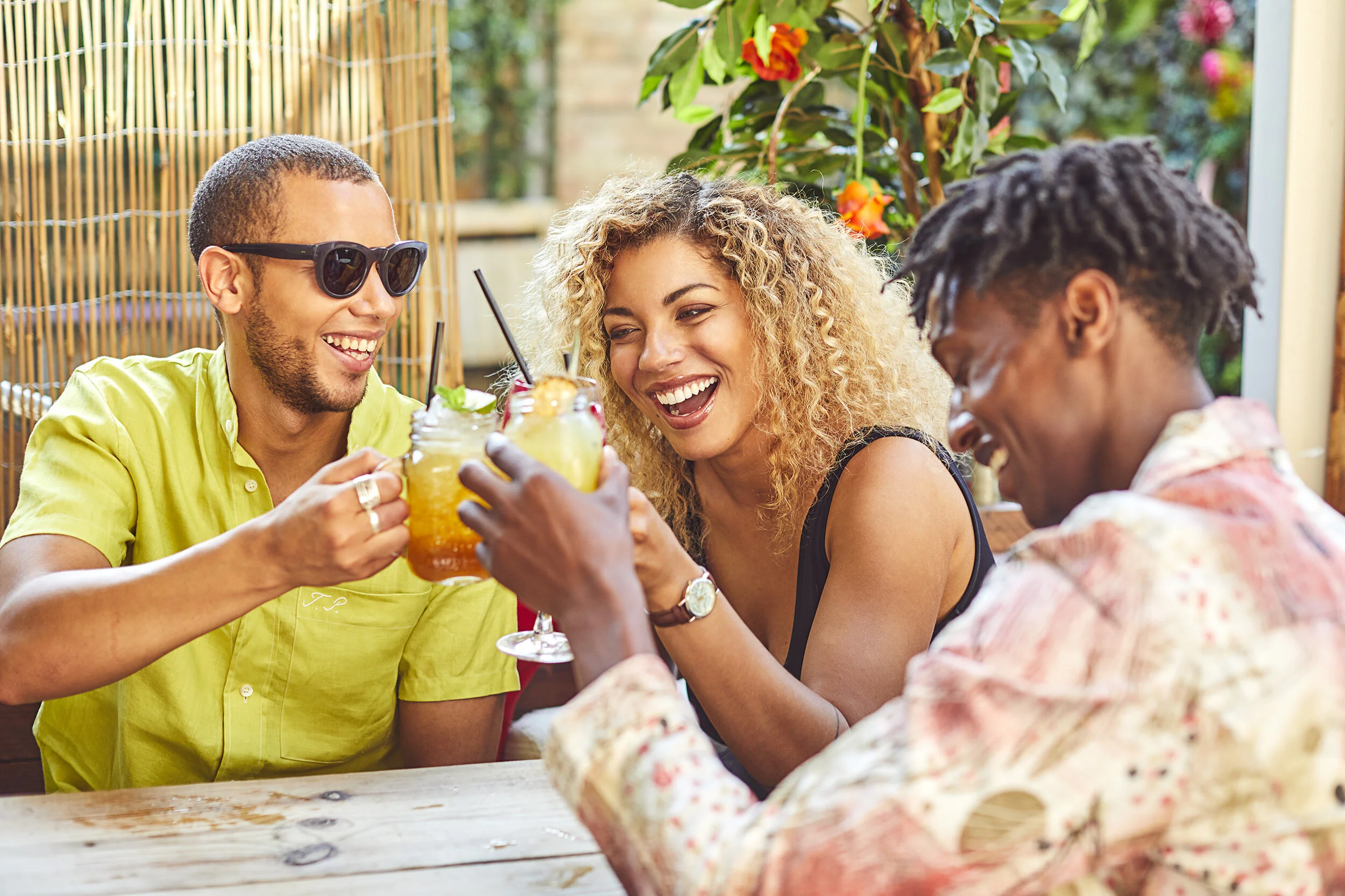 Three friends enjoying drinks and toasting at an outdoor table, with a colorful and lush garden in the background.