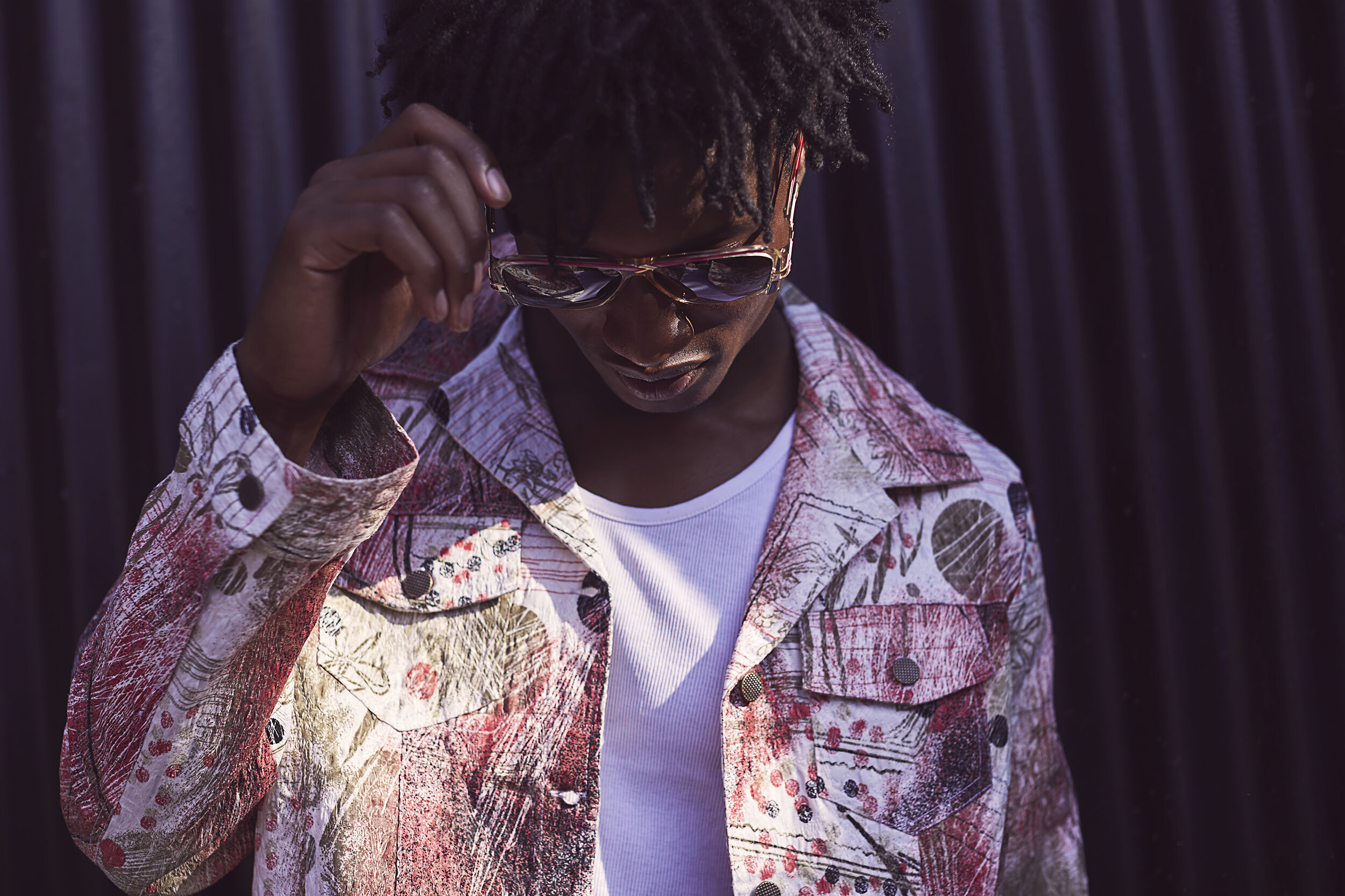 A young man with dark skin and dreadlocks wears reflective sunglasses, a colorful patterned denim jacket, and a white t-shirt, adjusting his sunglasses against a dark background.