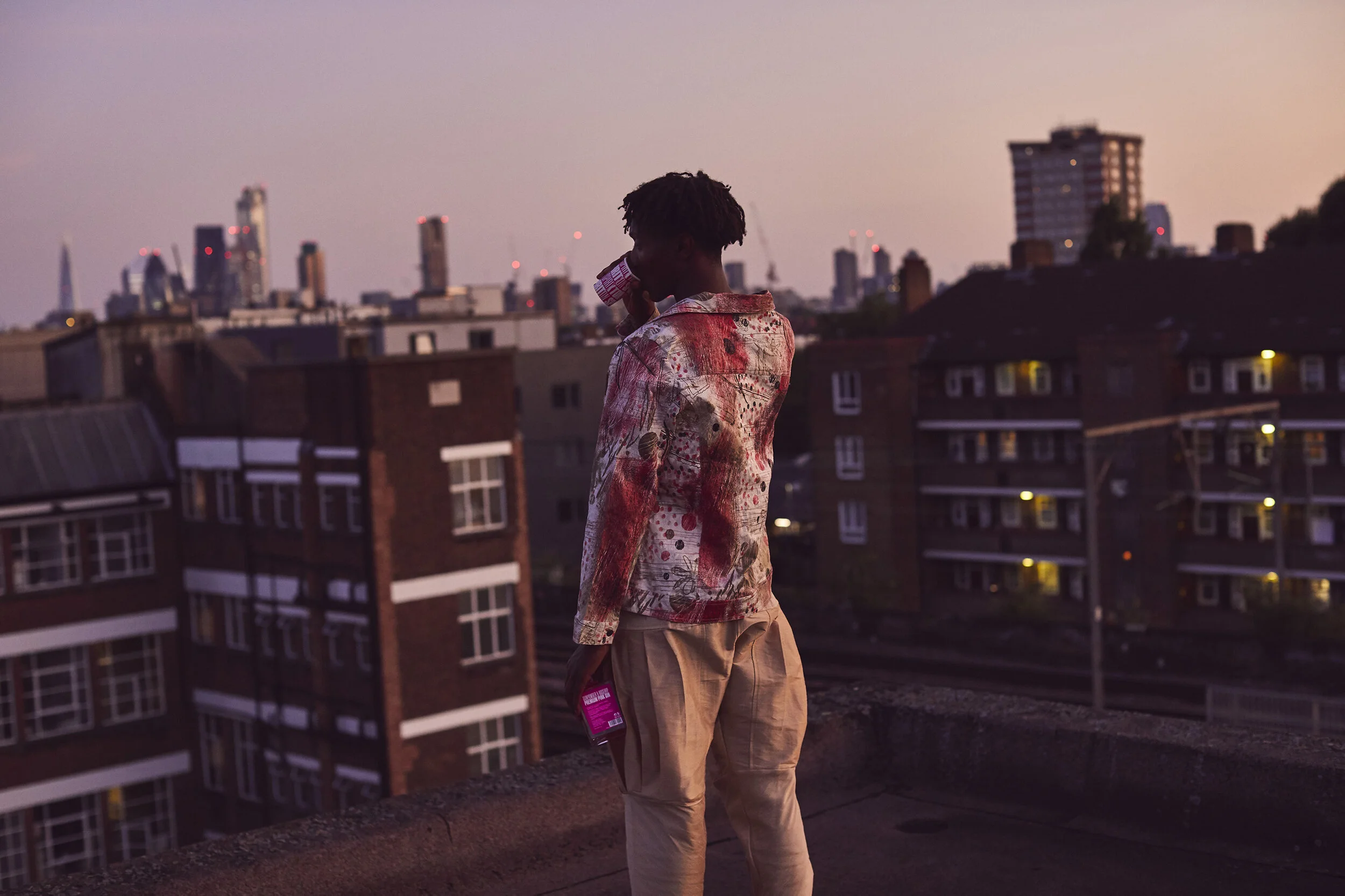 A young man stands on a rooftop at dusk, talking on a cell phone, with the city skyline and high-rise buildings in the background.