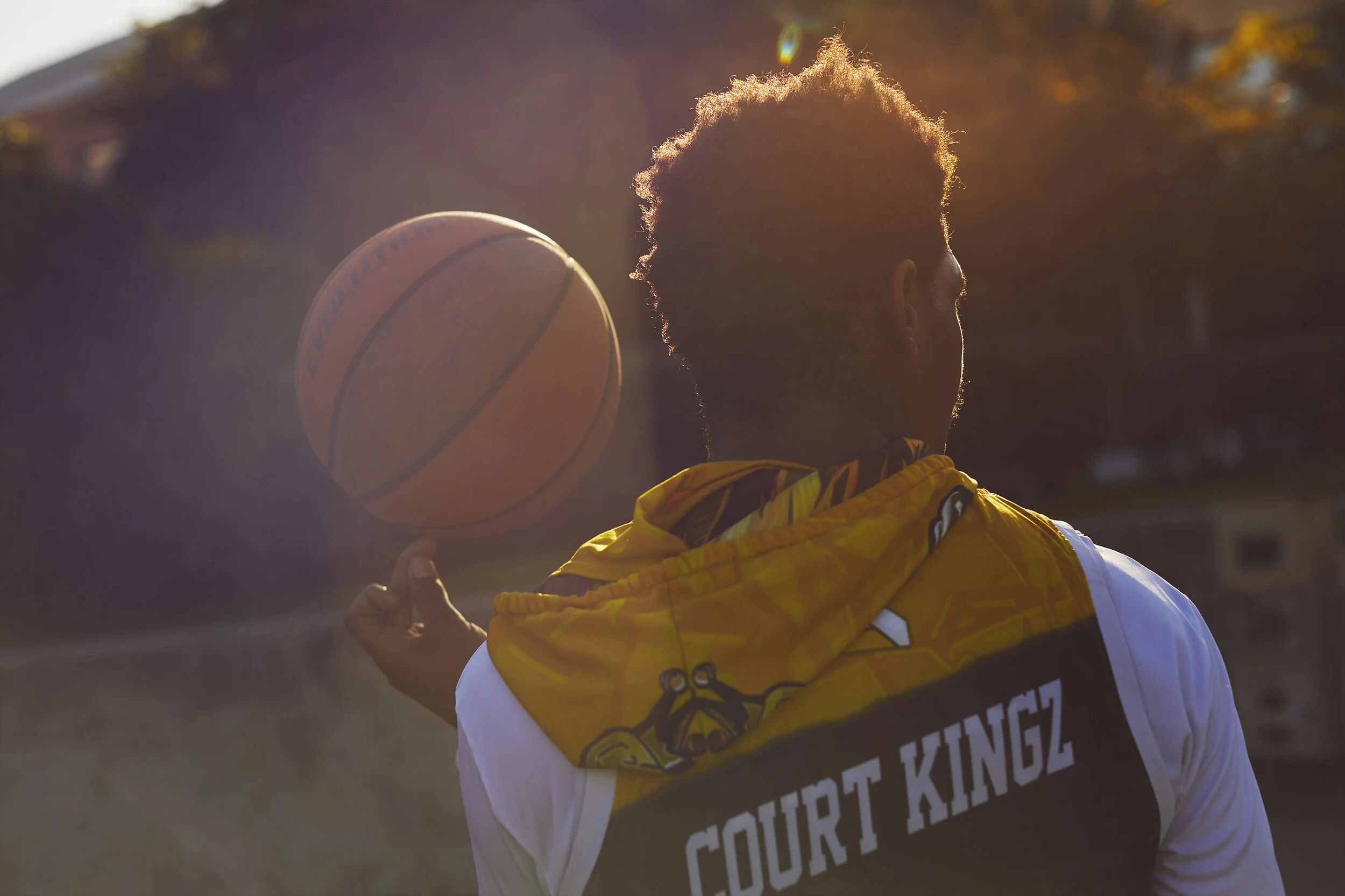 A person wearing a white shirt and a yellow jacket with a hood, with the words 'COURT KINGZ' on the back, dribbling a basketball outdoors during sunset.