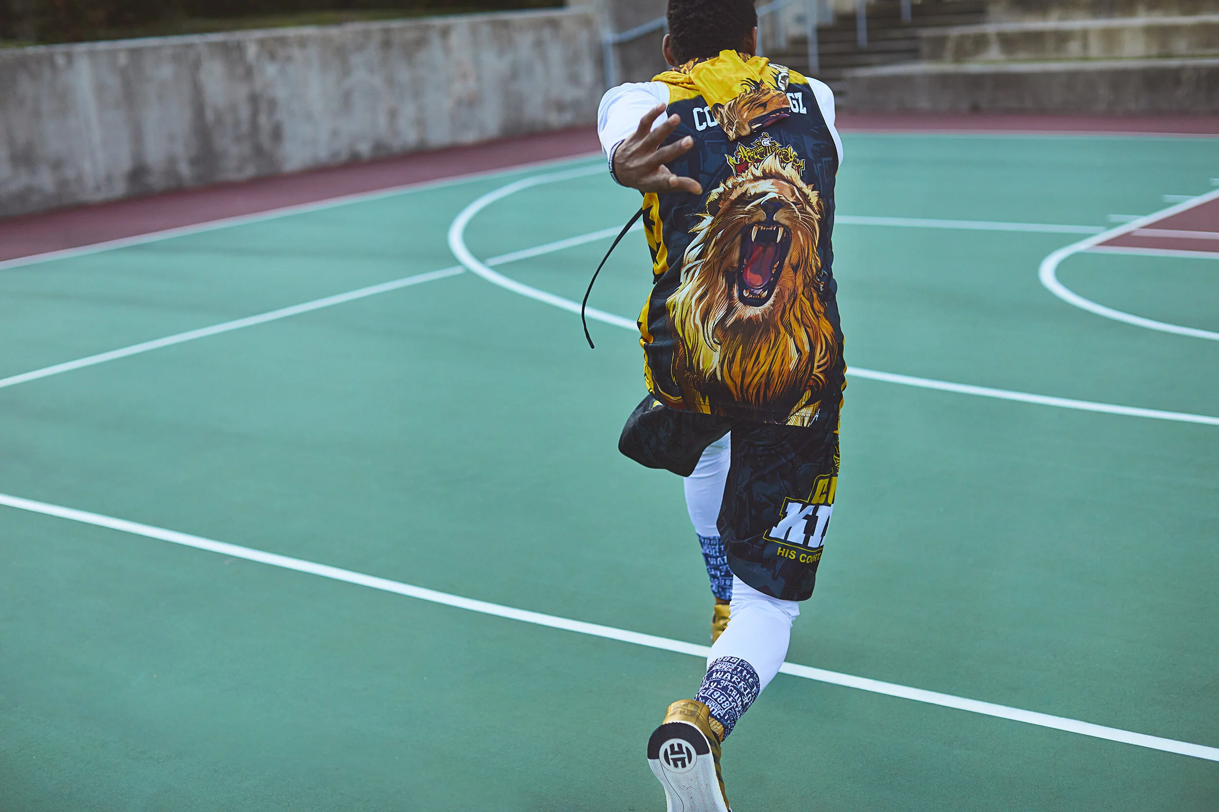 A person running on an outdoor sports court wearing a colorful outfit with a lion design on the back, making a gesture with one hand.