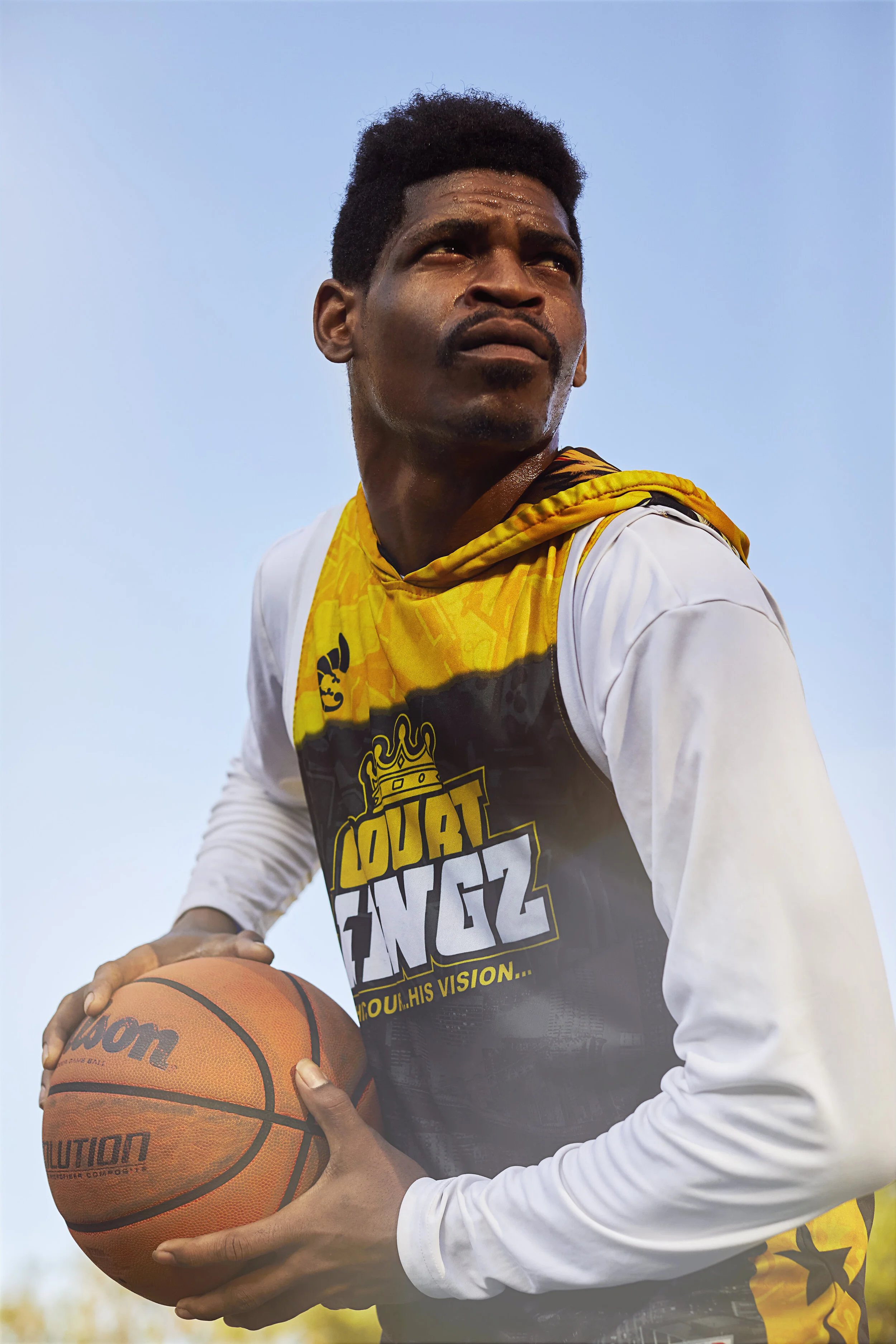 A male basketball player holding a basketball, wearing a white and yellow jersey, looking up against a clear blue sky.