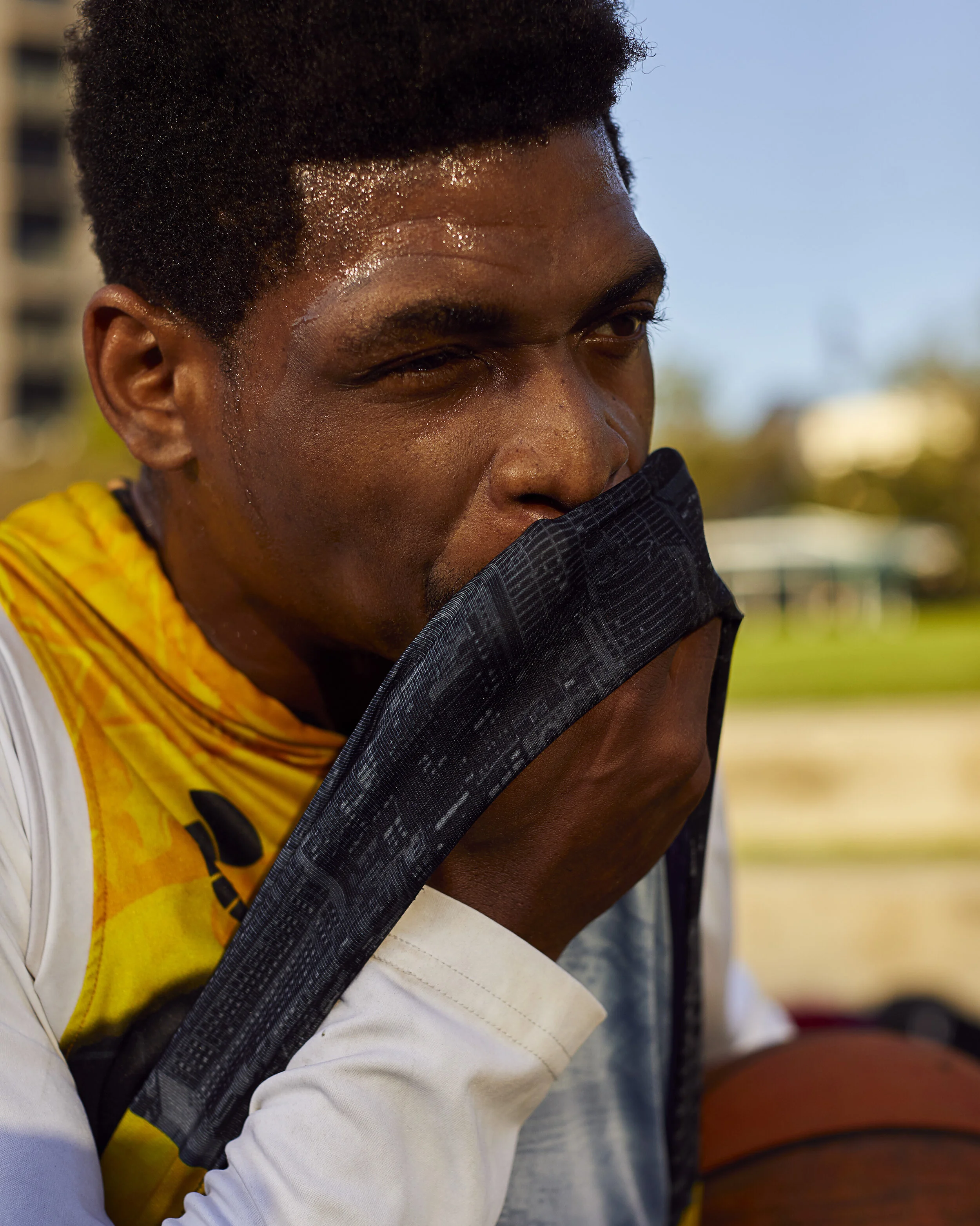 A young man sweating, holding a black towel over his mouth, outdoors during the daytime.