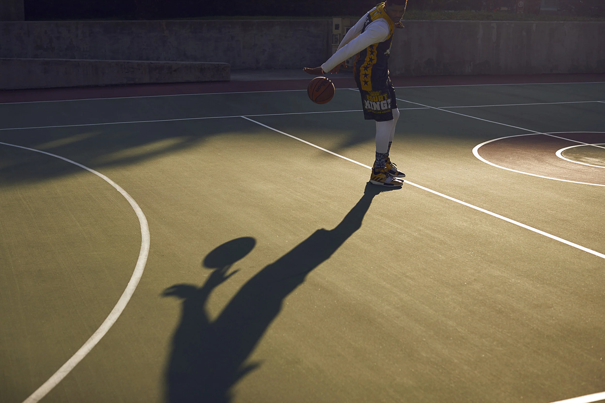 A young man in a basketball uniform is standing on an outdoor basketball court, holding a basketball, with his shadow cast on the ground.