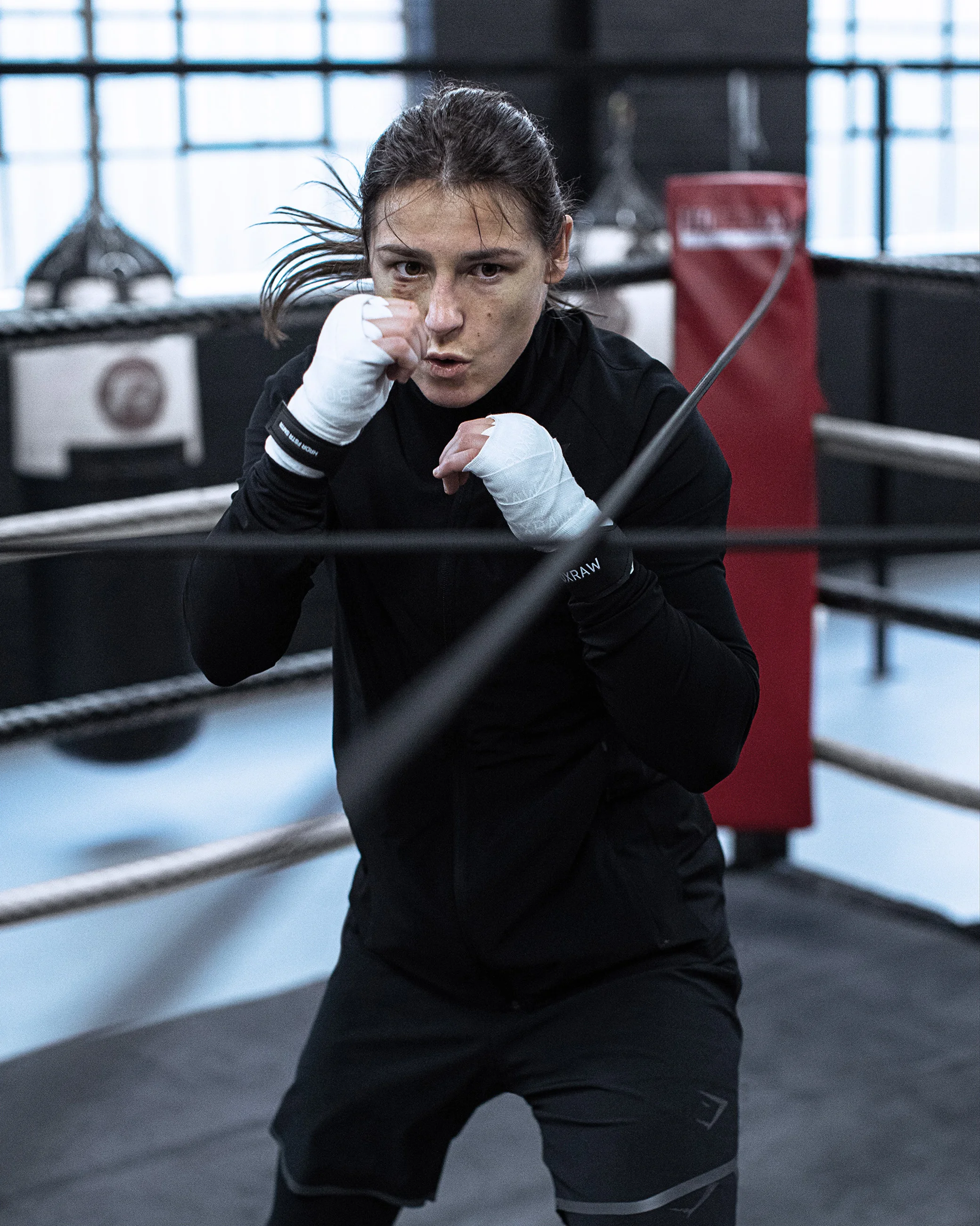 A female boxer in a training gym, standing in a fighting stance with fists up, ready to spar or practice.