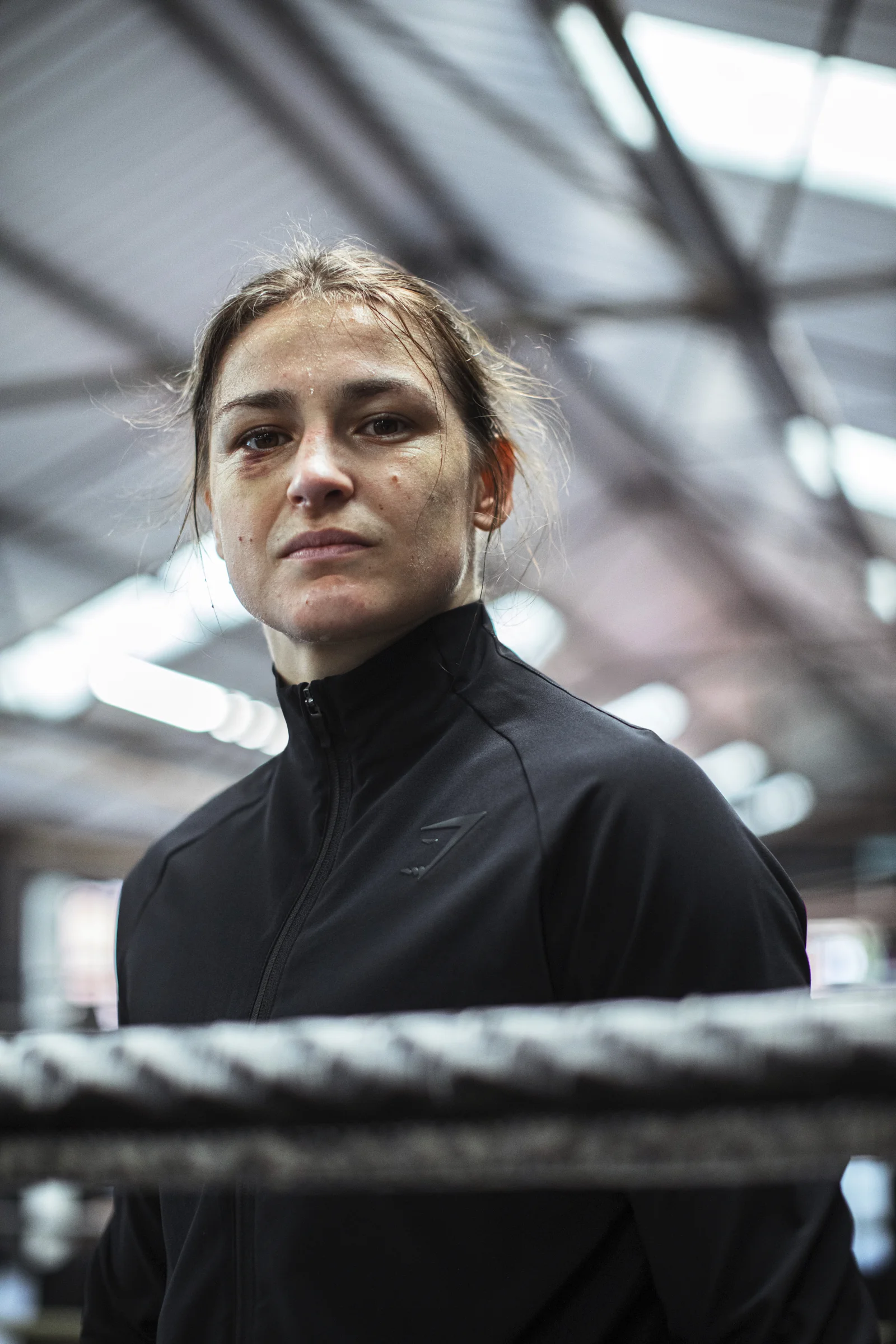 A young woman dressed in a black athletic jacket with a zip, standing inside a gym or sports facility with a high ceiling and metal beams visible in the background.