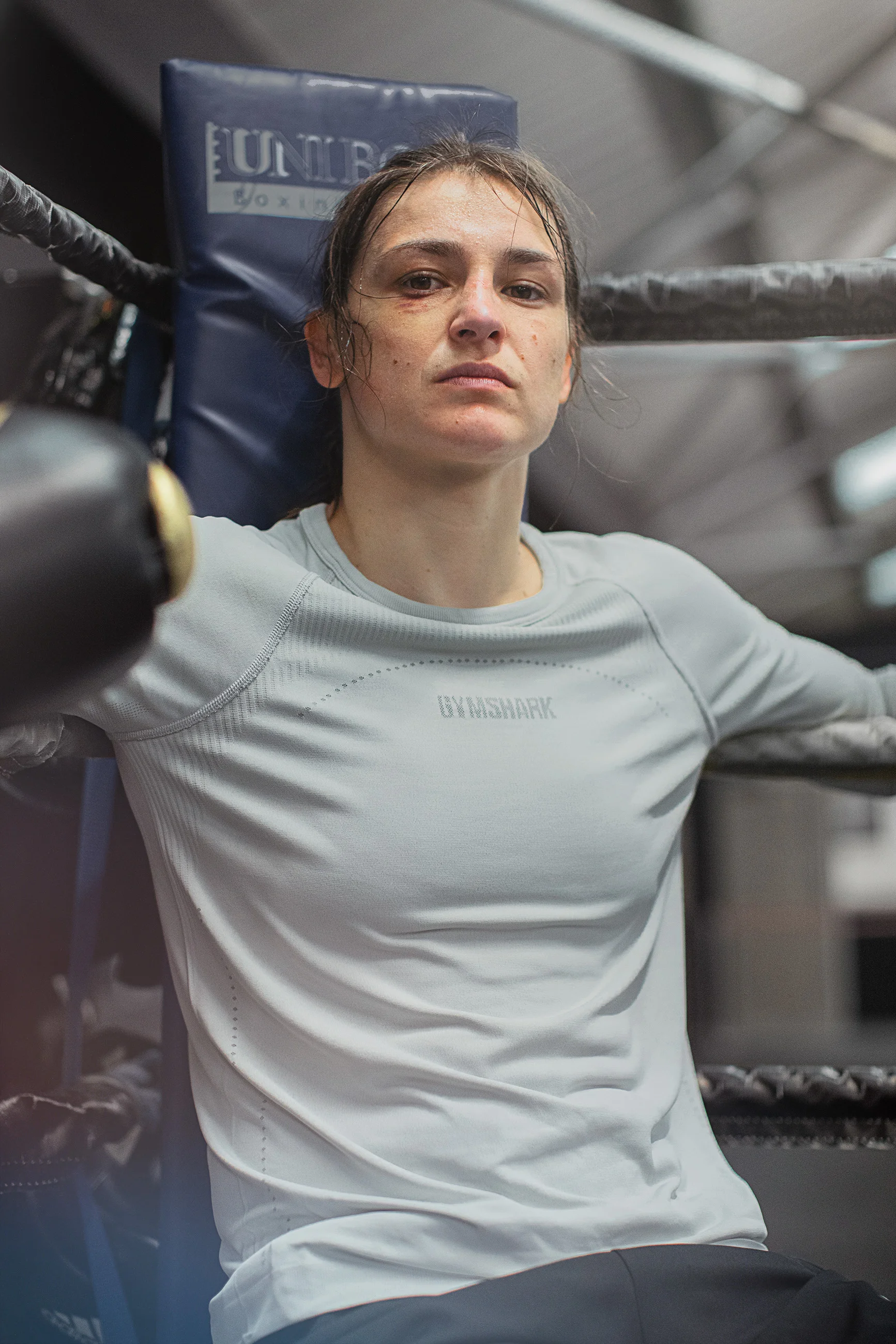 A female boxer rests with her back against the corner of a boxing ring, wearing a light gray athletic shirt and looking serious.