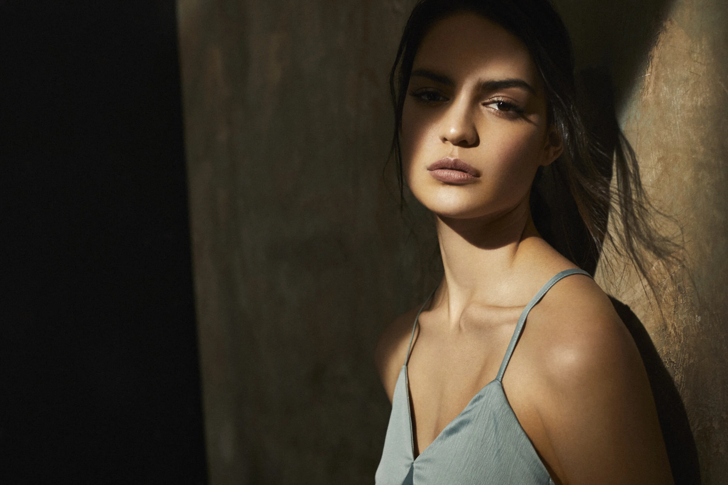 A woman with dark hair and neutral makeup looking at the camera, wearing a light blue spaghetti strap top, standing against a textured wall with a shadowed side.