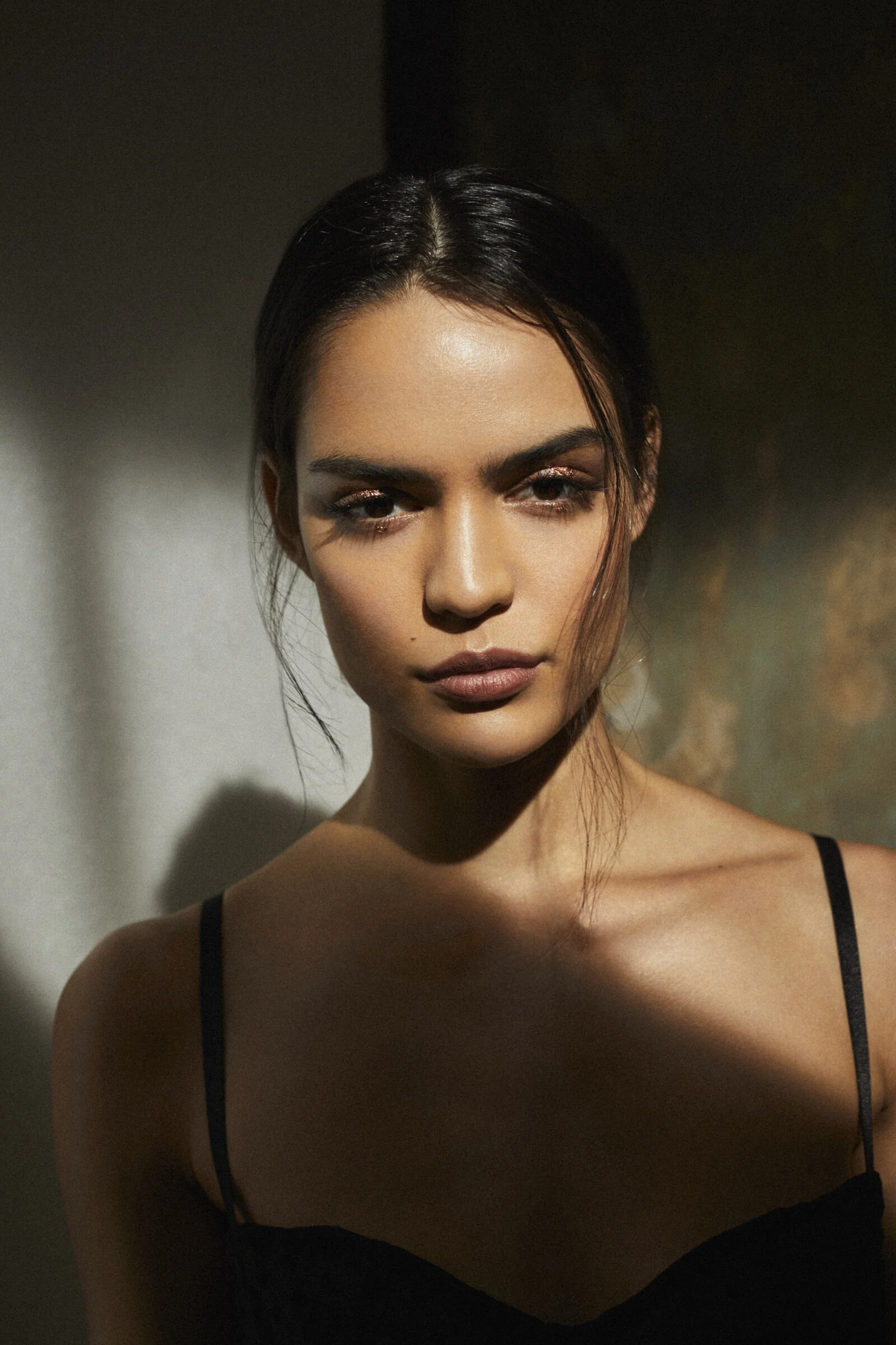 A woman with dark hair, wearing makeup, and a black spaghetti strap top, standing in front of a textured wall with shadows cast across her face and shoulders.
