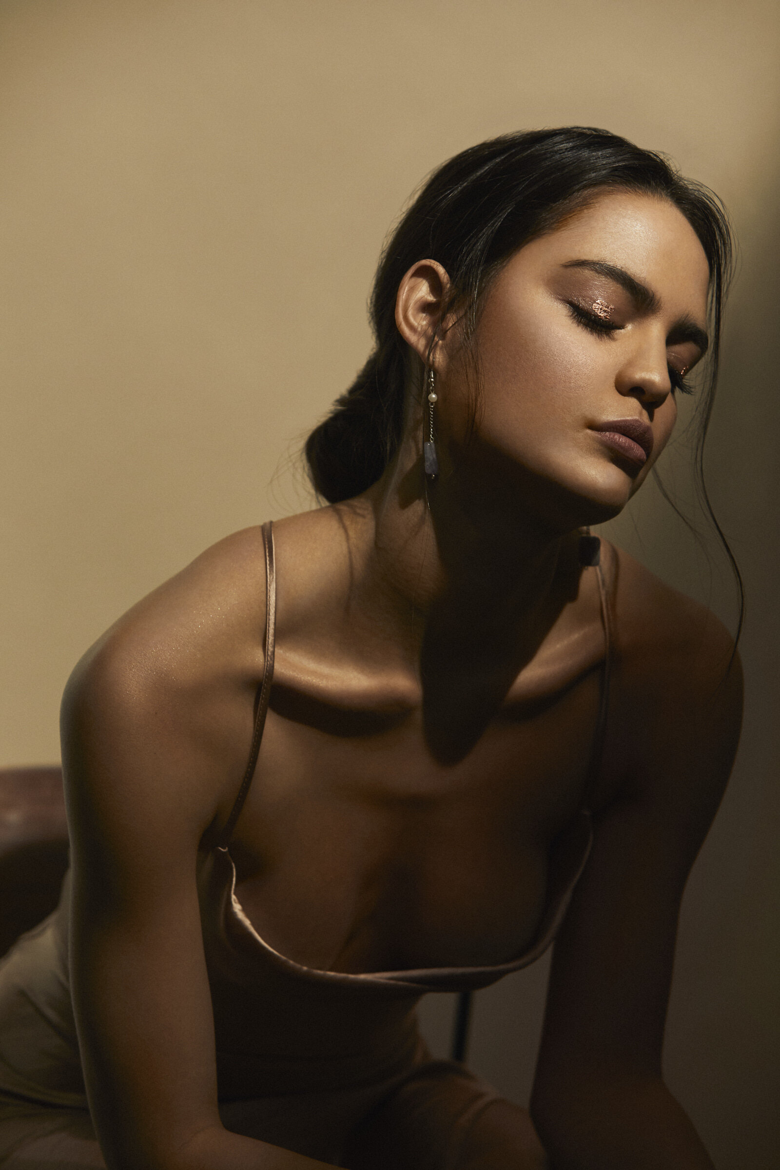 A woman with dark hair, closed eyes, and makeup, wearing a satin camisole and earrings, sitting against a plain beige background.