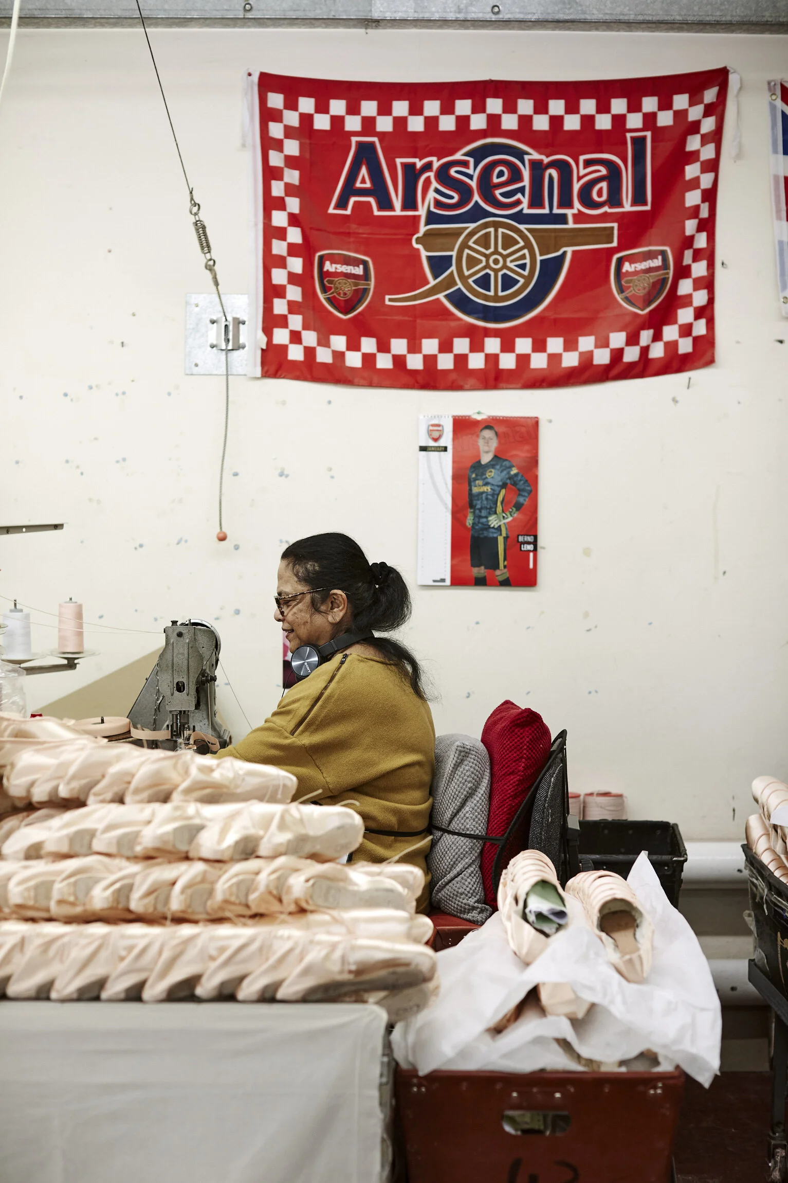 A woman sitting at a worktable with fabric, sewing shoes in a factory. A red Arsenal football banner and a poster of a soccer player are on the wall behind her.