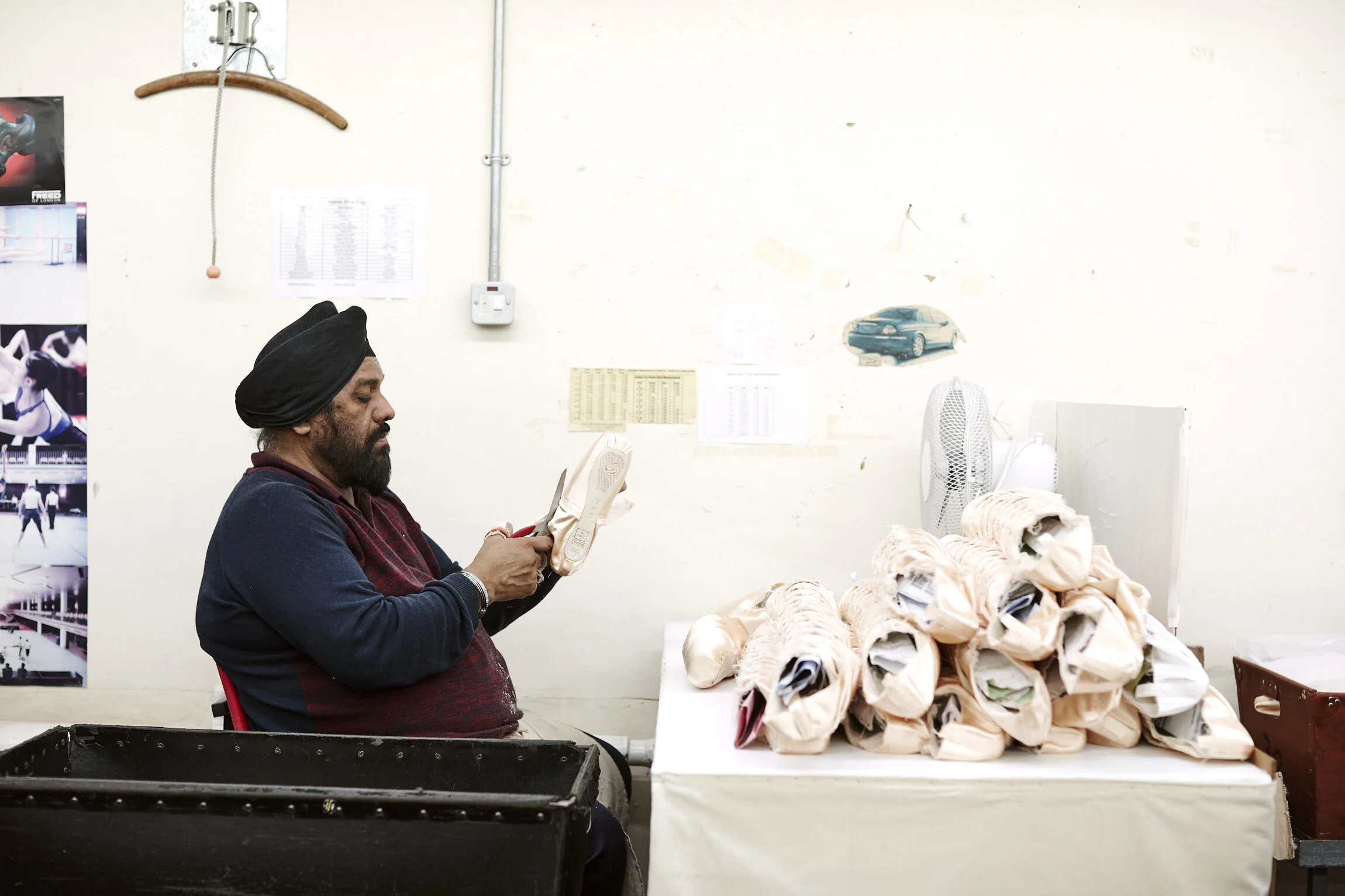 A man with a beard and turban sitting at a table, holding a cloth item. On the table are numerous wrapped items, possibly shoes or cloth bundles. The background includes posters, a fan, and a wall with papers and small pictures.
