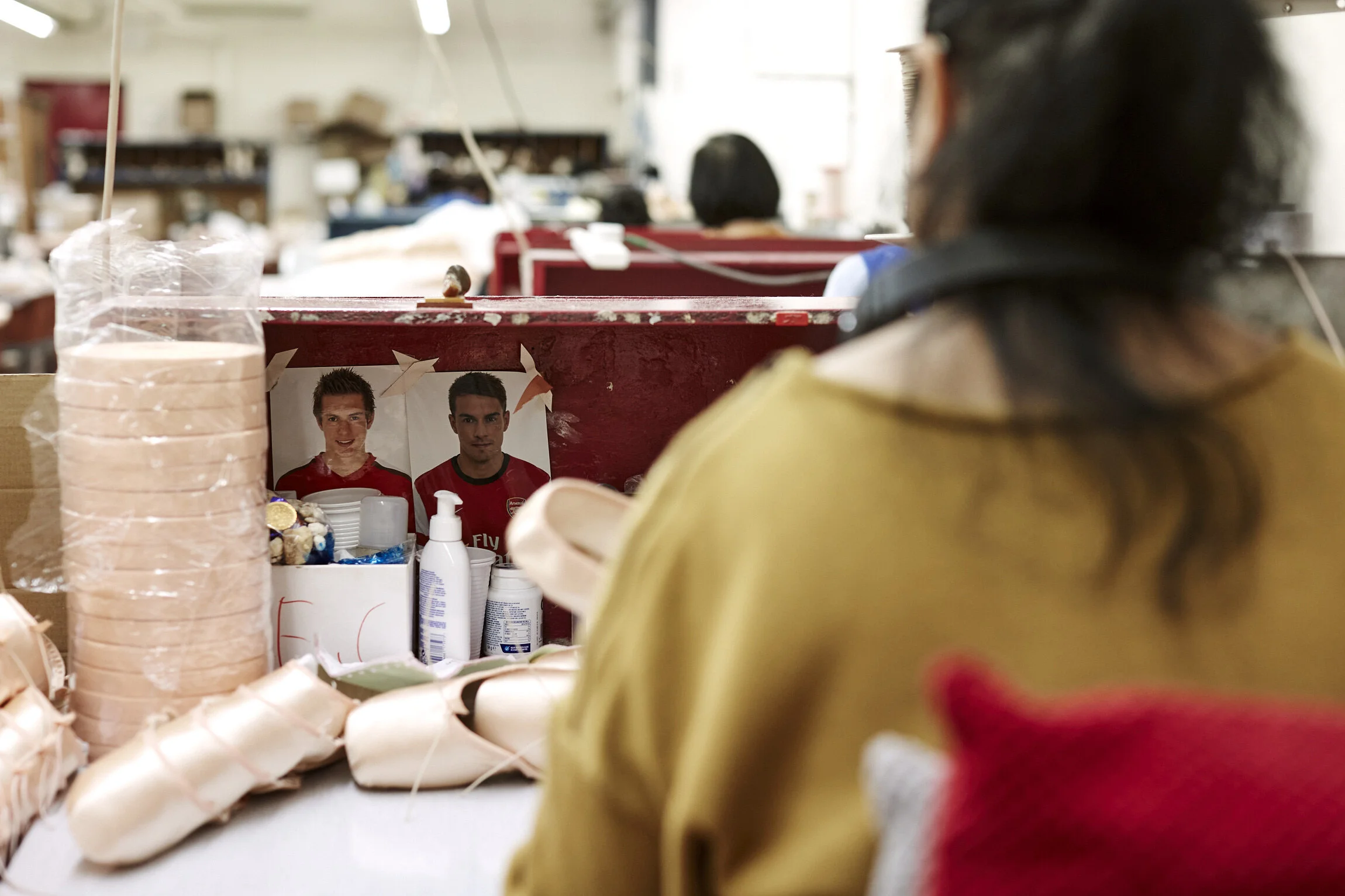 A woman with dark hair and sunglasses sitting at a table, with a vigil or memorial setup in front of her featuring photos of two soccer players in red jerseys, candles, and flowers.