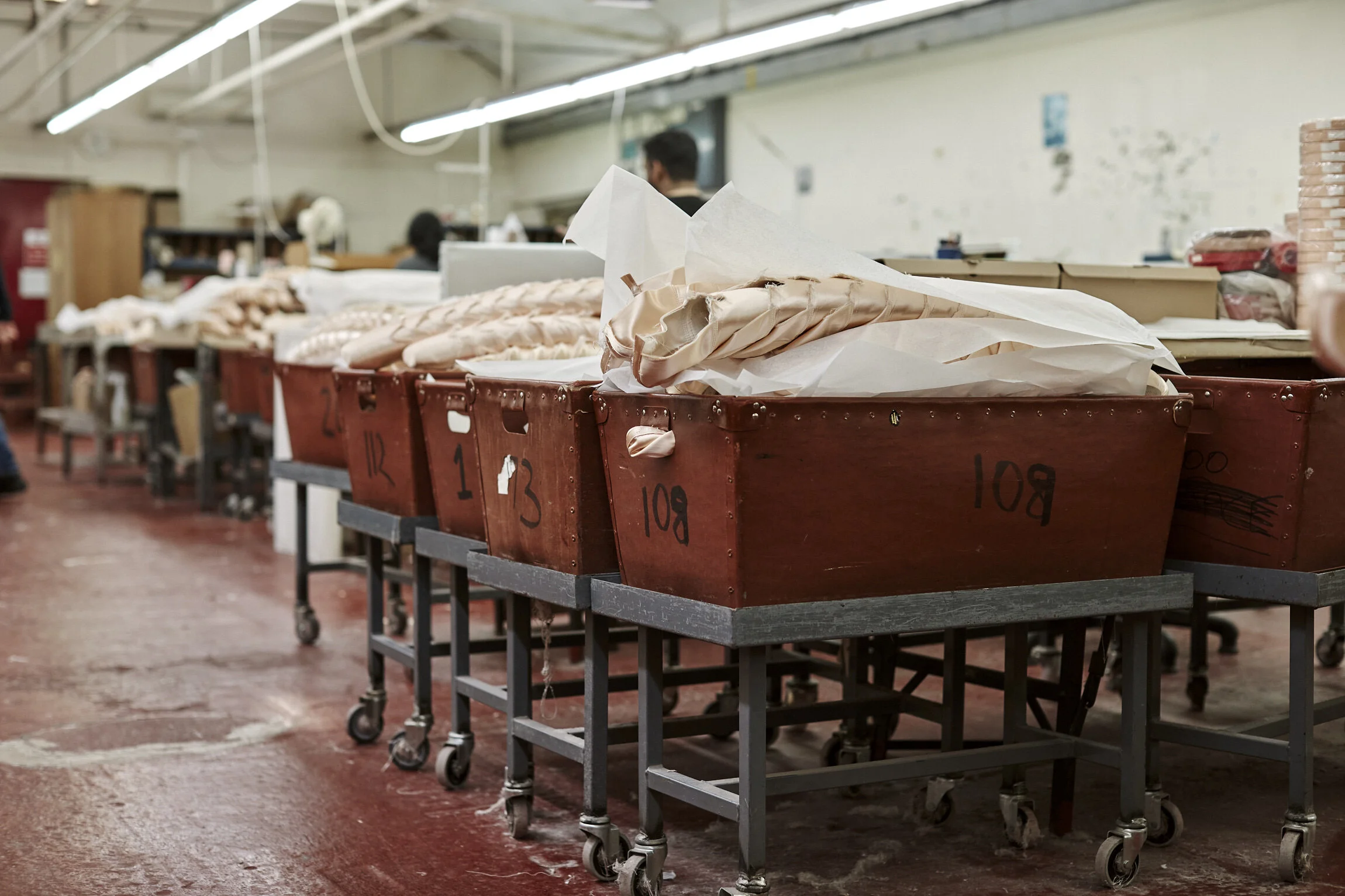 Industrial workspace with red numbered carts on wheels, containing fabric or soft materials, in a factory or workshop with metal tables and people working in the background.