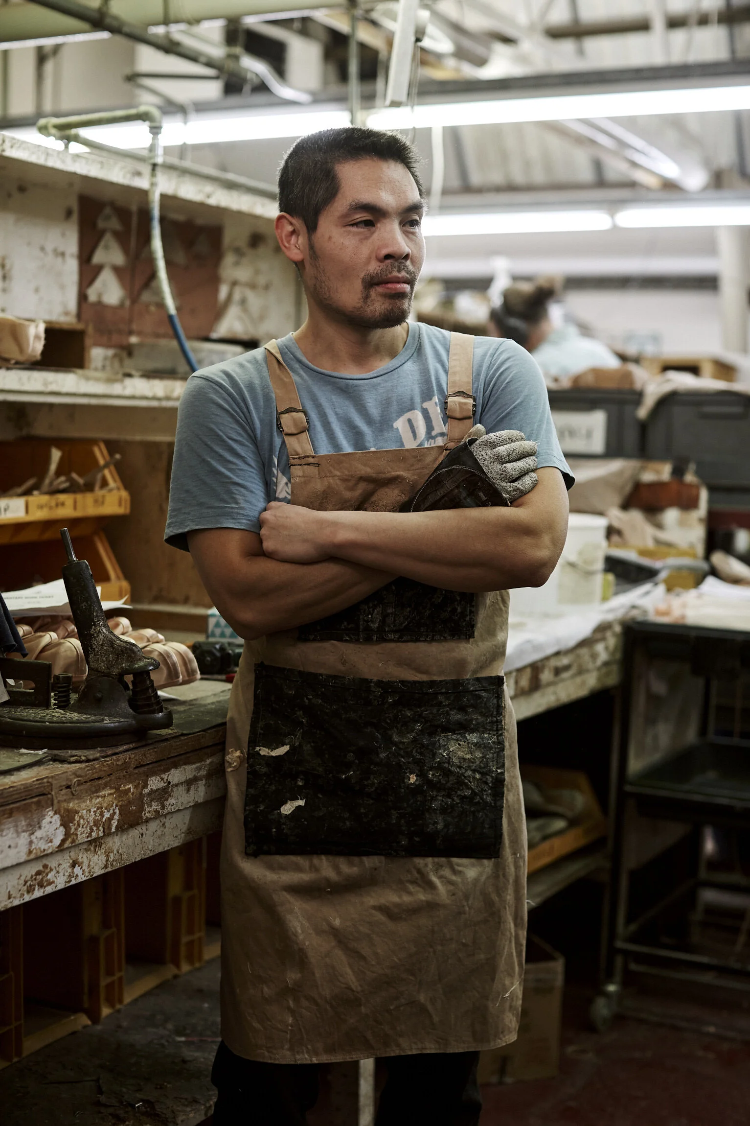 A man wearing a blue t-shirt and a worn apron, standing with arms crossed in a workshop or factory setting, surrounded by various tools and materials.
