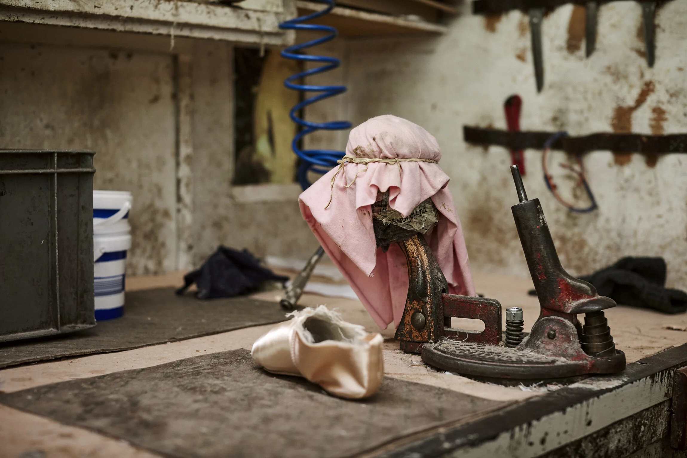 A dusty workbench with an old, rusty shoe repair tool, a pink cloth covering its top, a worn-out white ballet shoe, and various tools hanging on a peeling wall in the background.
