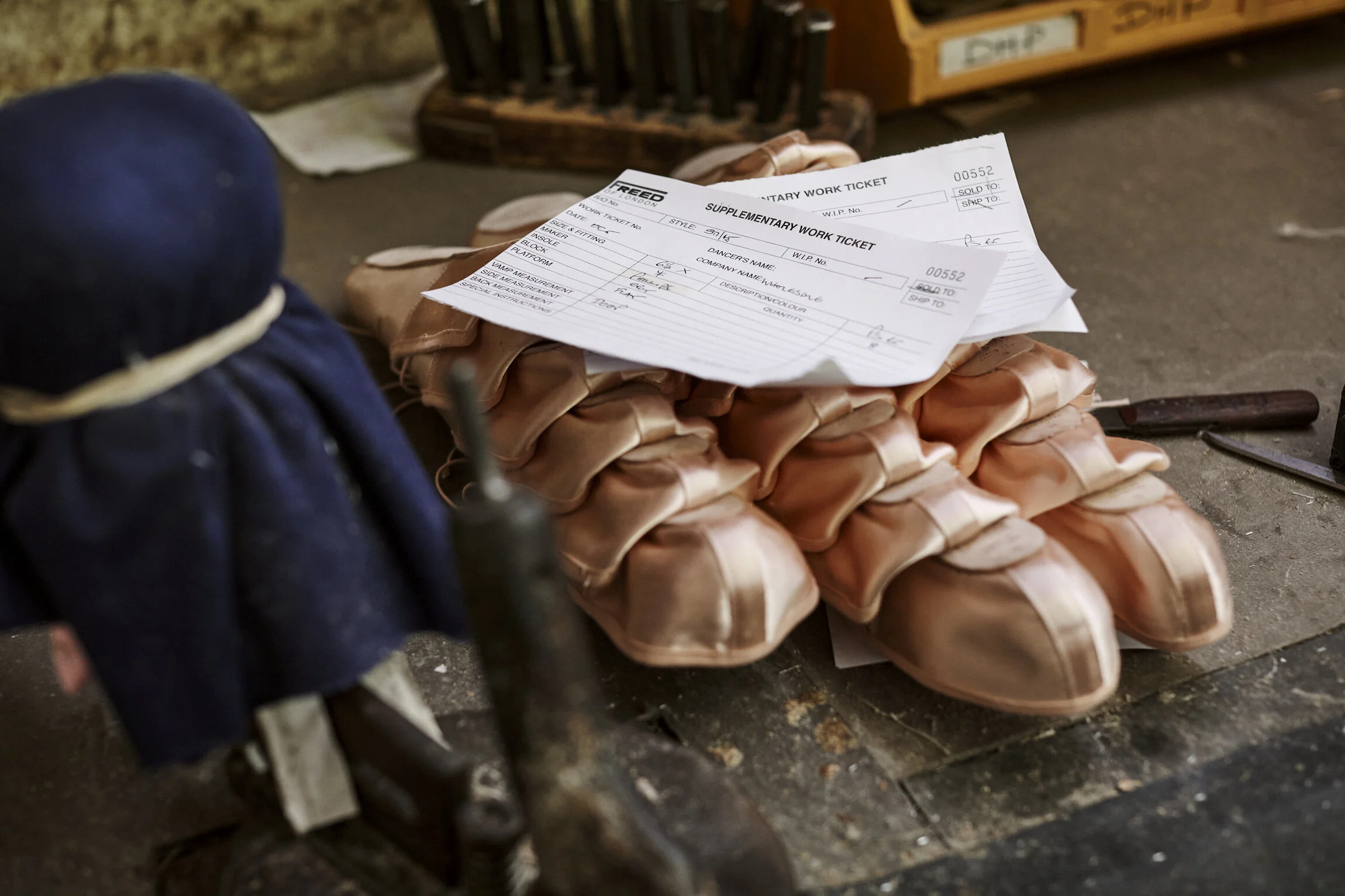 A pile of ballet pointe shoes with two supplementary work tickets placed on top, on a dusty black floor in a dance studio or backstage area.