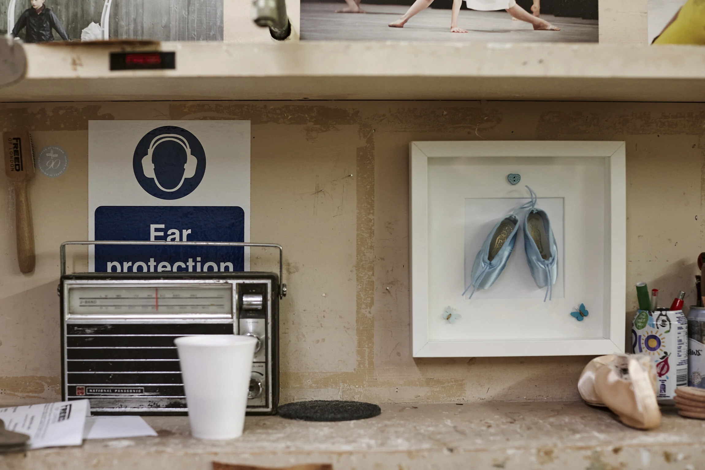 A shelf with a blue 'Ear protection' sign, an old radio, a white cup, a framed display of ballet shoes with butterfly decorations, and various objects including a shoe, a tube of cream, and a few papers.