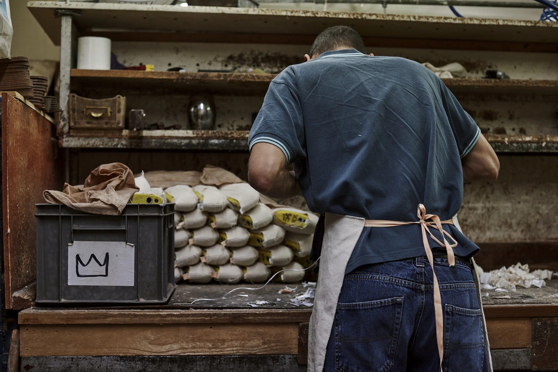 Person with dark hair, wearing a blue polo shirt and an apron, working at a woodworking or craft table in a workshop with stacks of small bags, tools, and shelves in the background.