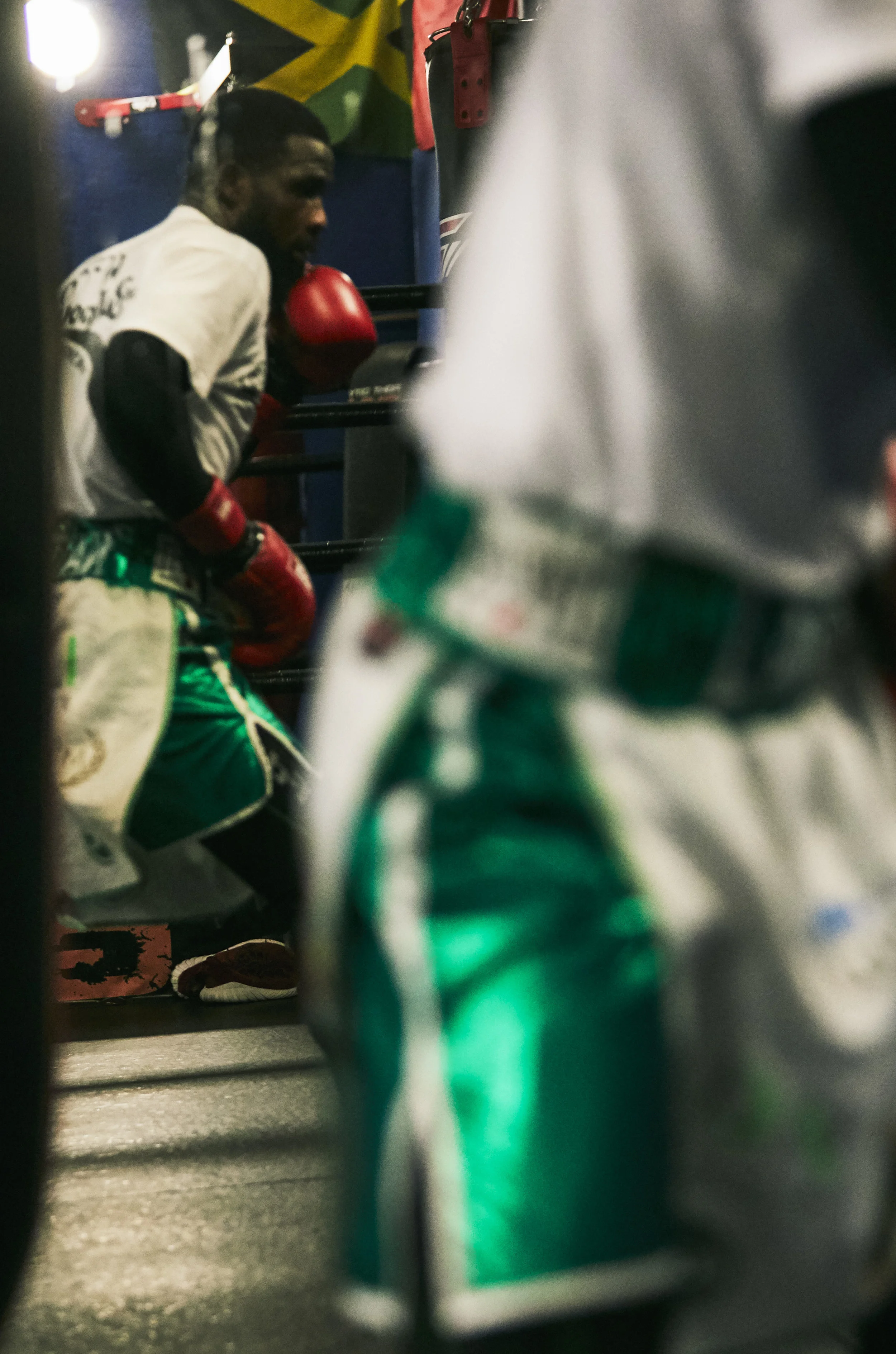 A male boxer wearing red gloves and green shorts in a boxing gym, with a Jamaican flag hanging in the background, seen through a gap in the equipment.